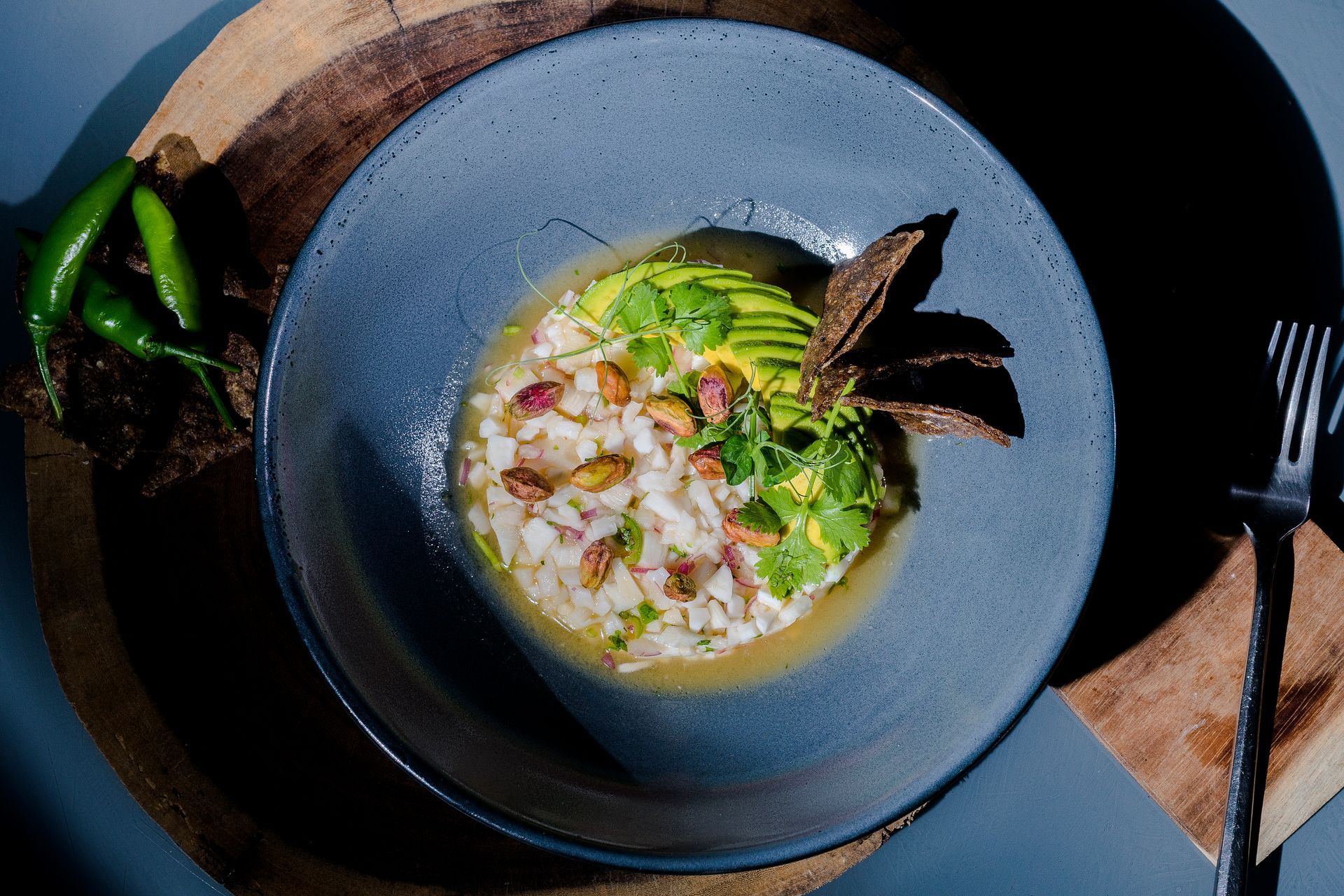 Primer plano de ceviche en un recipiente azul con aguacate, pistachos, cilantro y chips de tortilla.