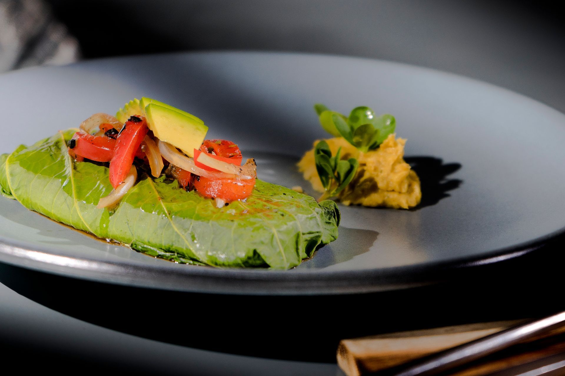 Un plato con un wrap de col rizada cubierto con aguacate y tomates, acompañado de salsa amarilla.