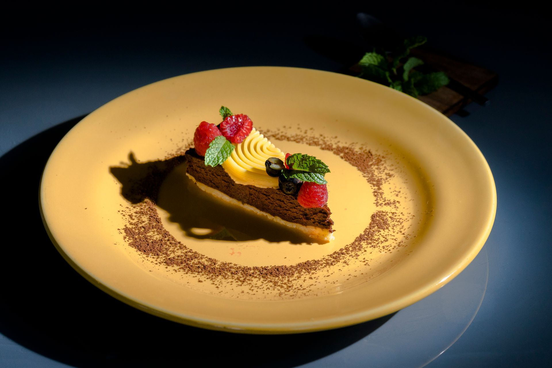 Rebanada de pastel de chocolate en un plato amarillo, decorada con frutos rojos, crema y menta. Espolvoreada con cacao.