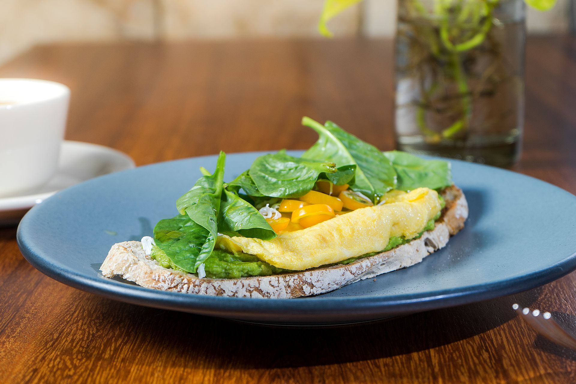 Tortilla con espinacas y mango sobre tostada, servida en un plato azul, con una taza de café y un jarrón con plantas verdes de fondo.