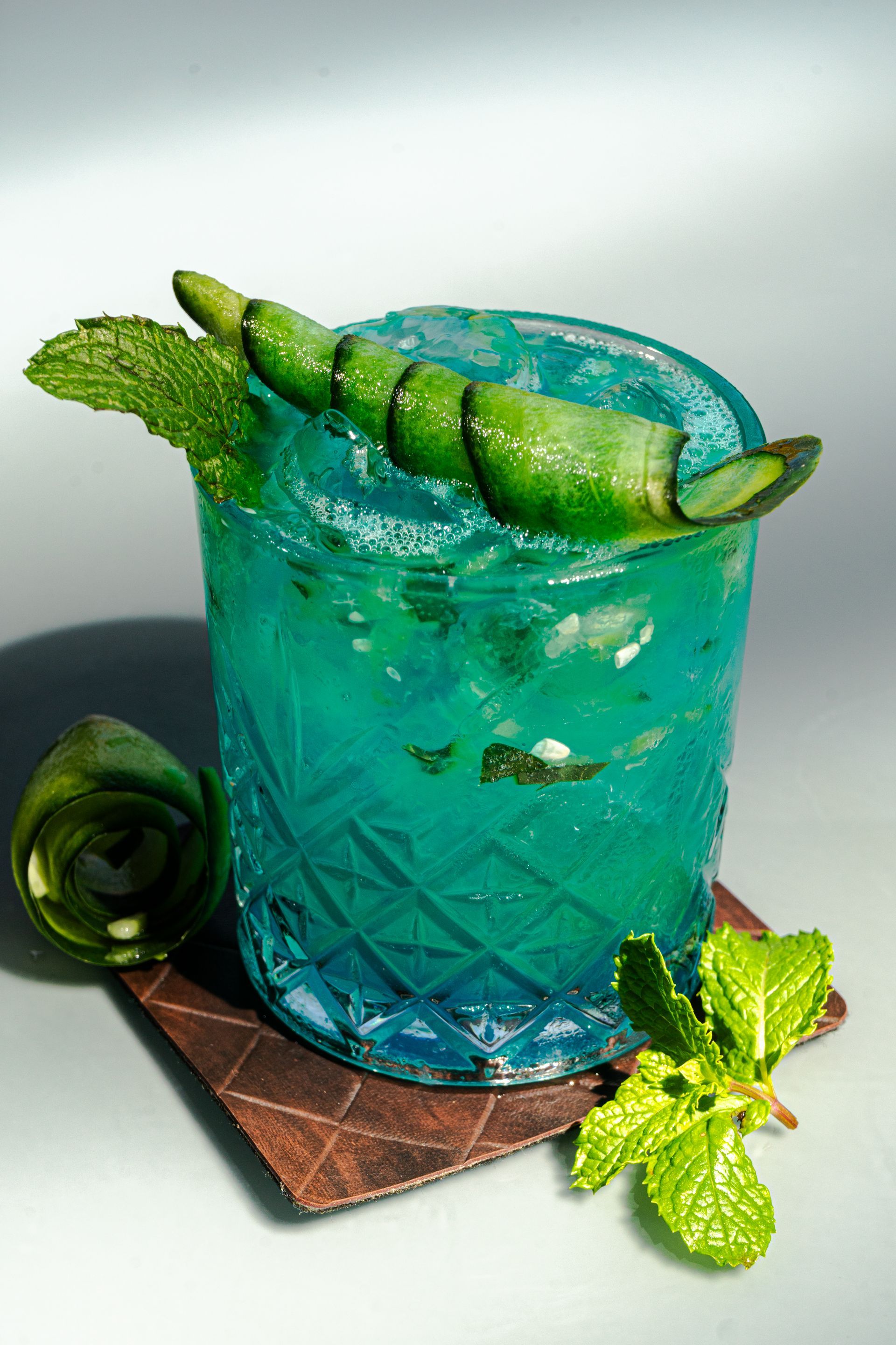 Cóctel azul en vaso texturizado, adornado con pepino y menta, sobre un posavasos.