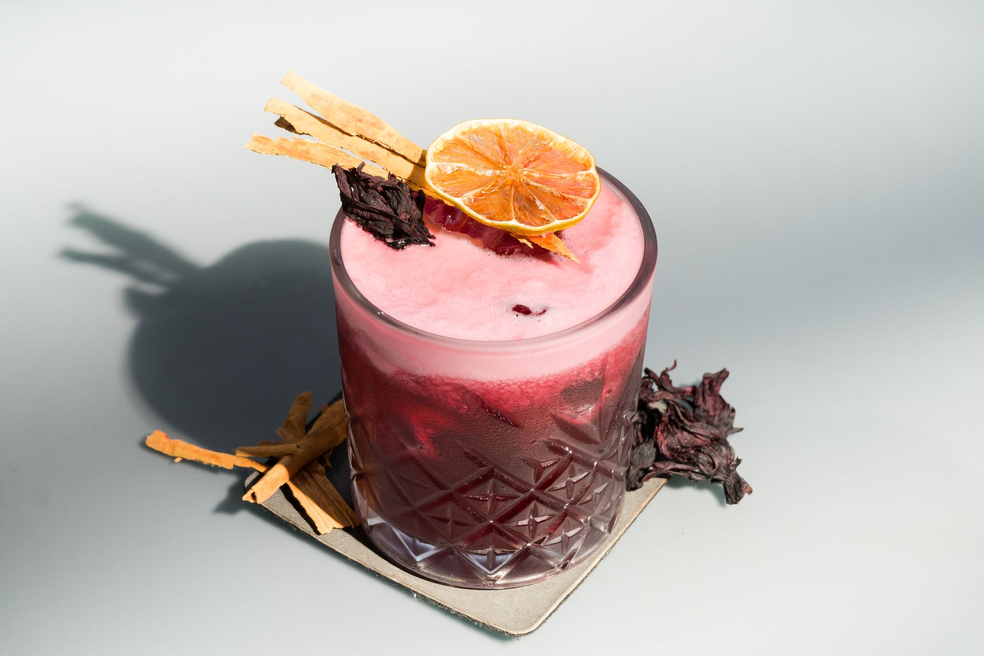 Cóctel de tono rosado, adornado con cítricos secos, ramas de canela y flores en una copa decorativa.