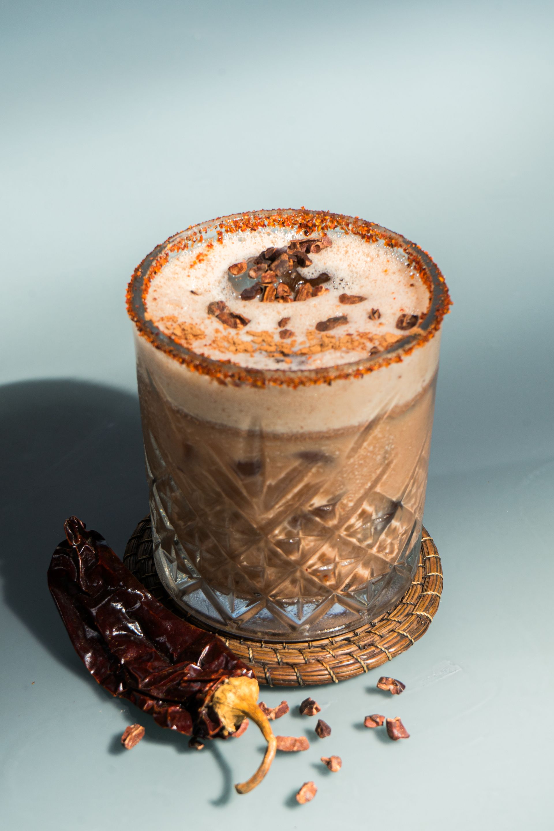 Cóctel de chocolate en copa estampada, guarnición de chile picante y fondo celeste.