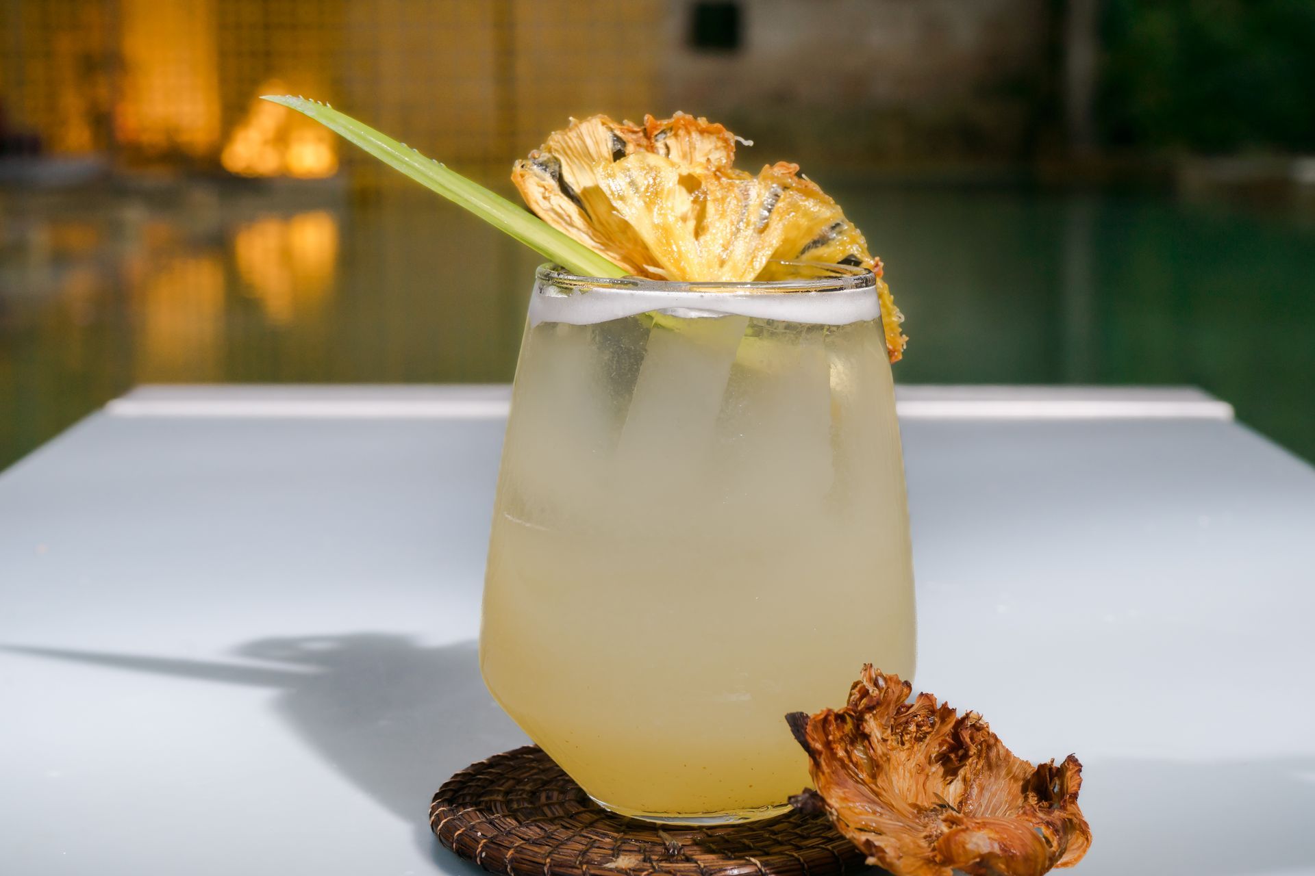 Cóctel en copa con decoración de piña y limoncillo sobre una superficie blanca, con una piscina al fondo.