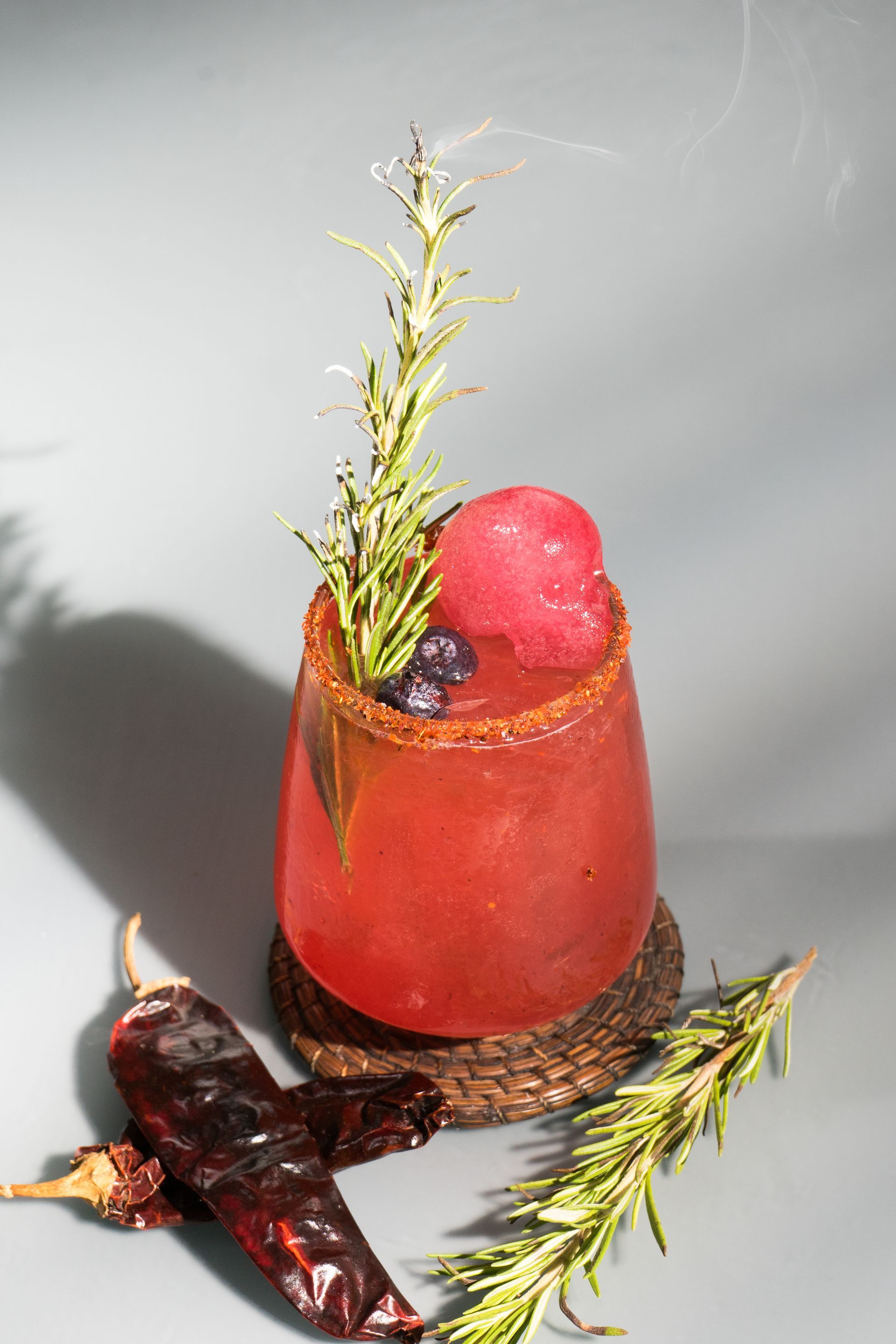Cóctel rojo con guarnición de chile picante y ramita de romero, sobre posavasos.