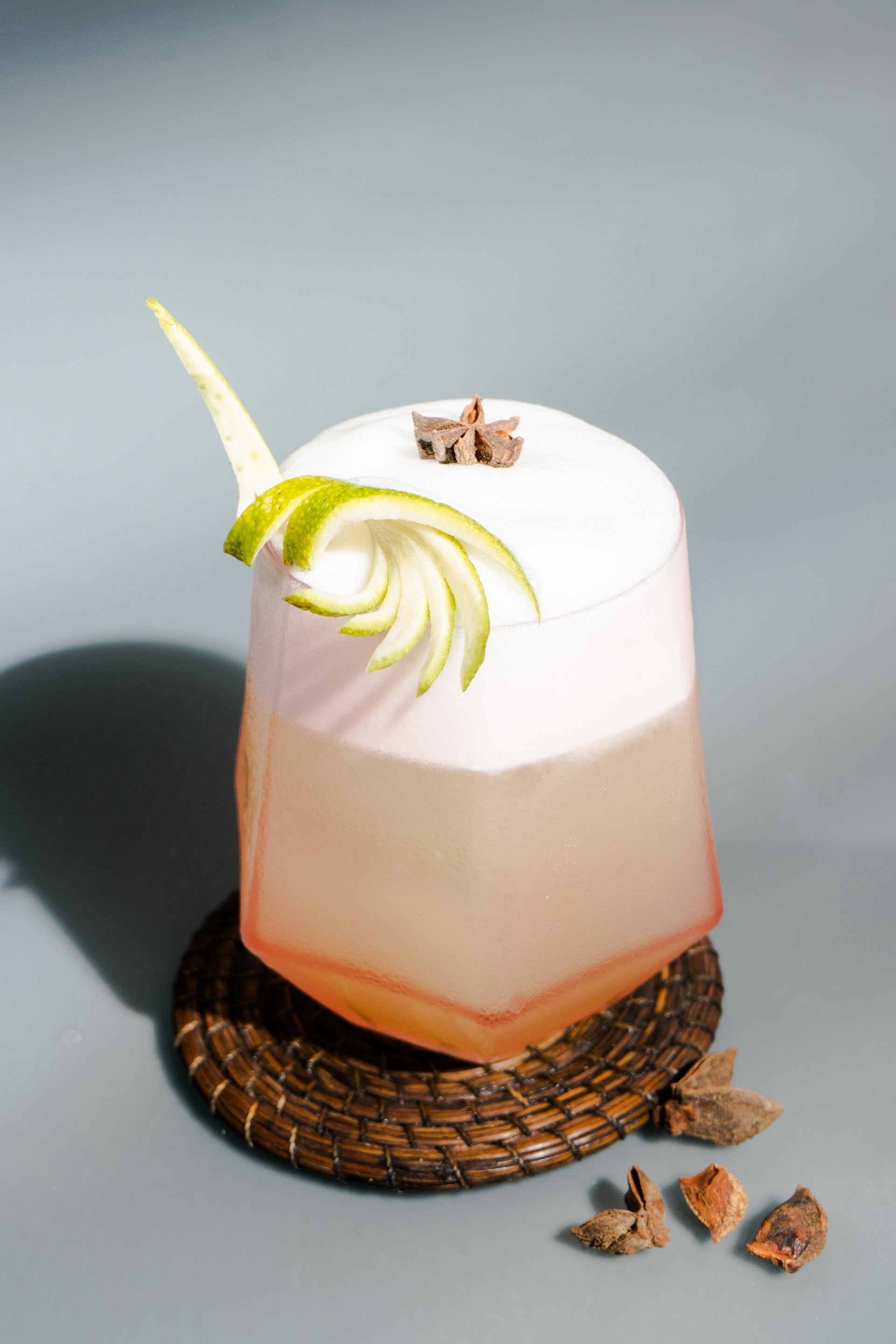 Cóctel en copa geométrica, con capa de espuma blanca en la parte superior y decoración de cáscara de lima sobre un posavasos tejido.
