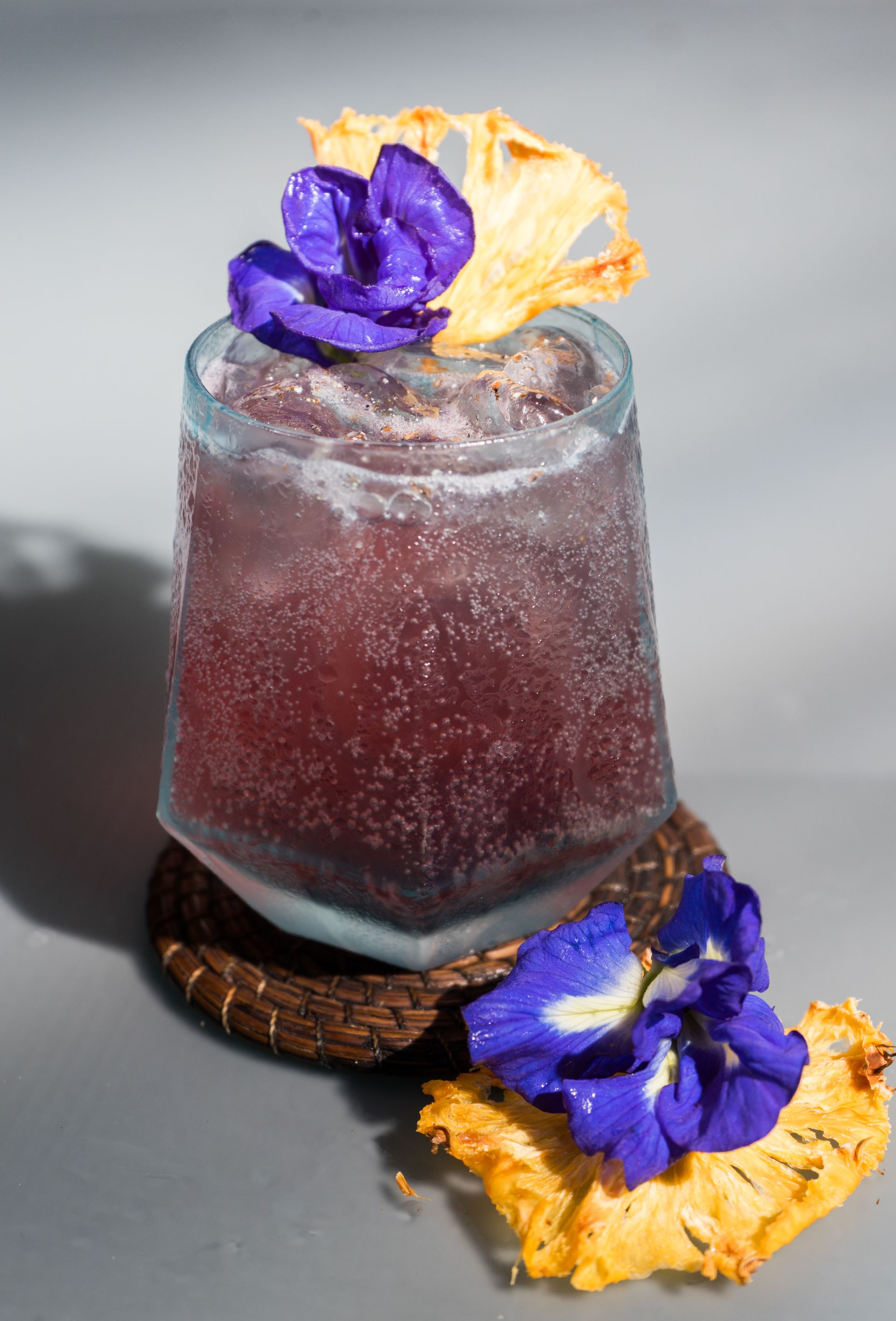 Cóctel morado con hielo en vaso, decorado con flores azules y piña.