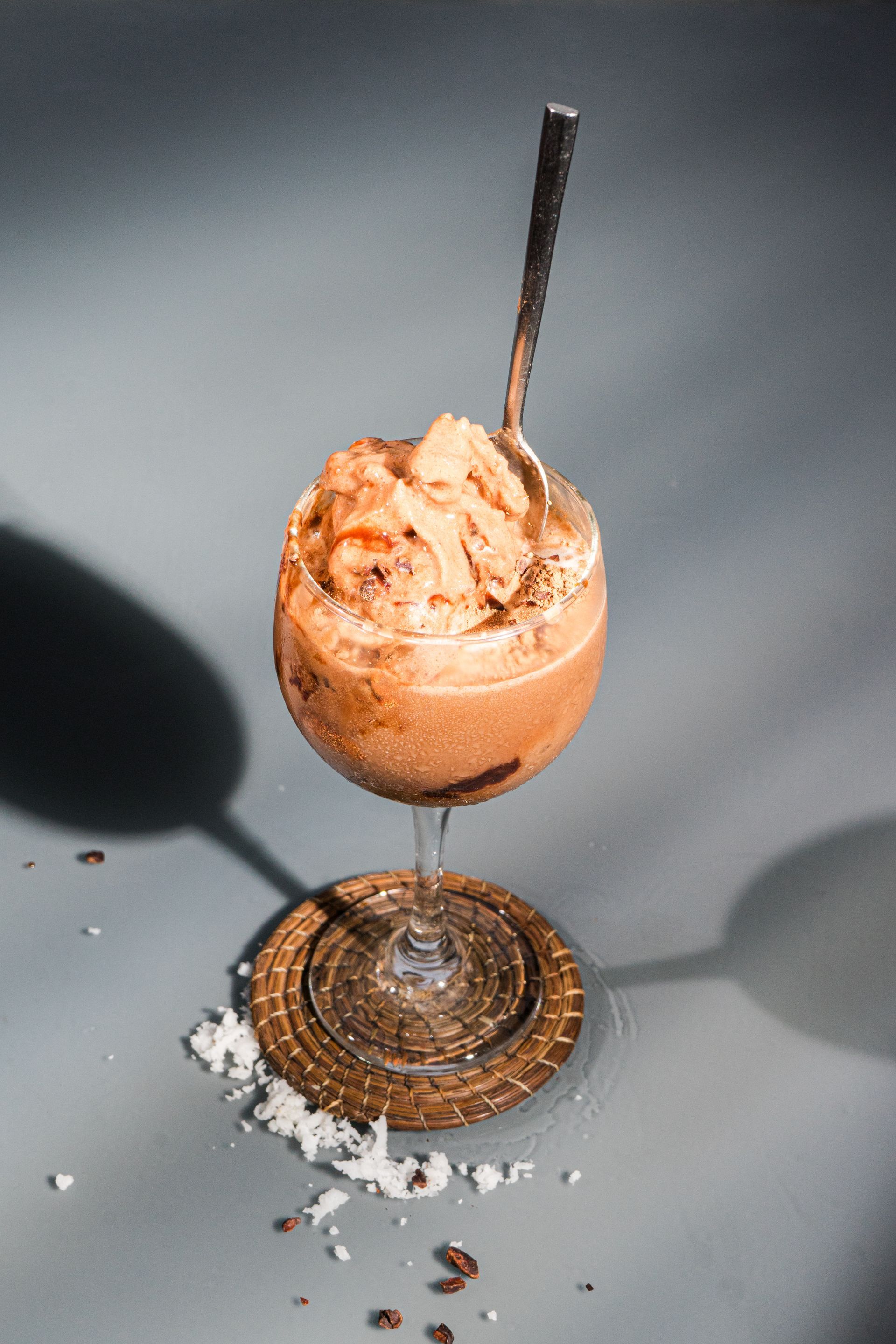 Postre de chocolate en copa con tallo y cuchara; sobre posavasos, fondo gris.