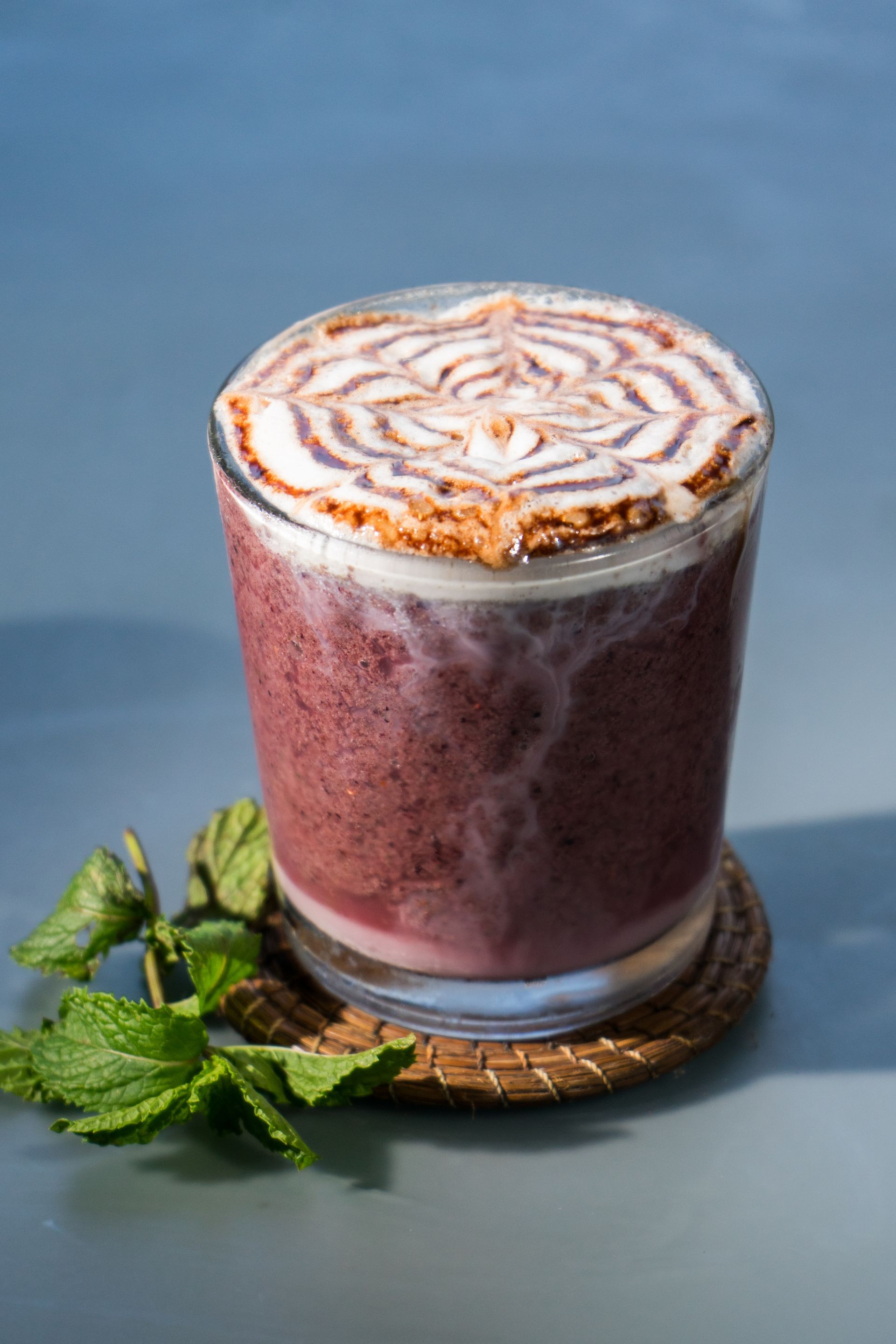 Batido morado con arte de espuma, sobre un posavasos tejido, ramita de menta.