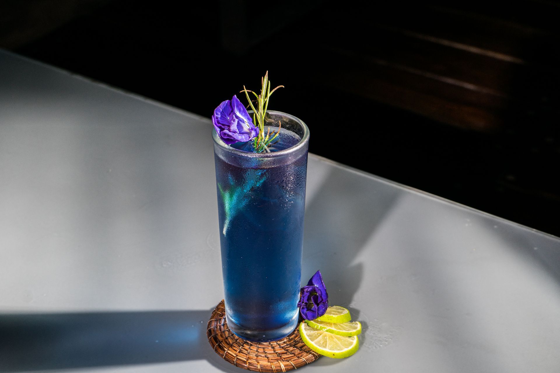 Cóctel azul en vaso alto, adornado con una flor y hierbas, sobre una barra con rodajas de limón.