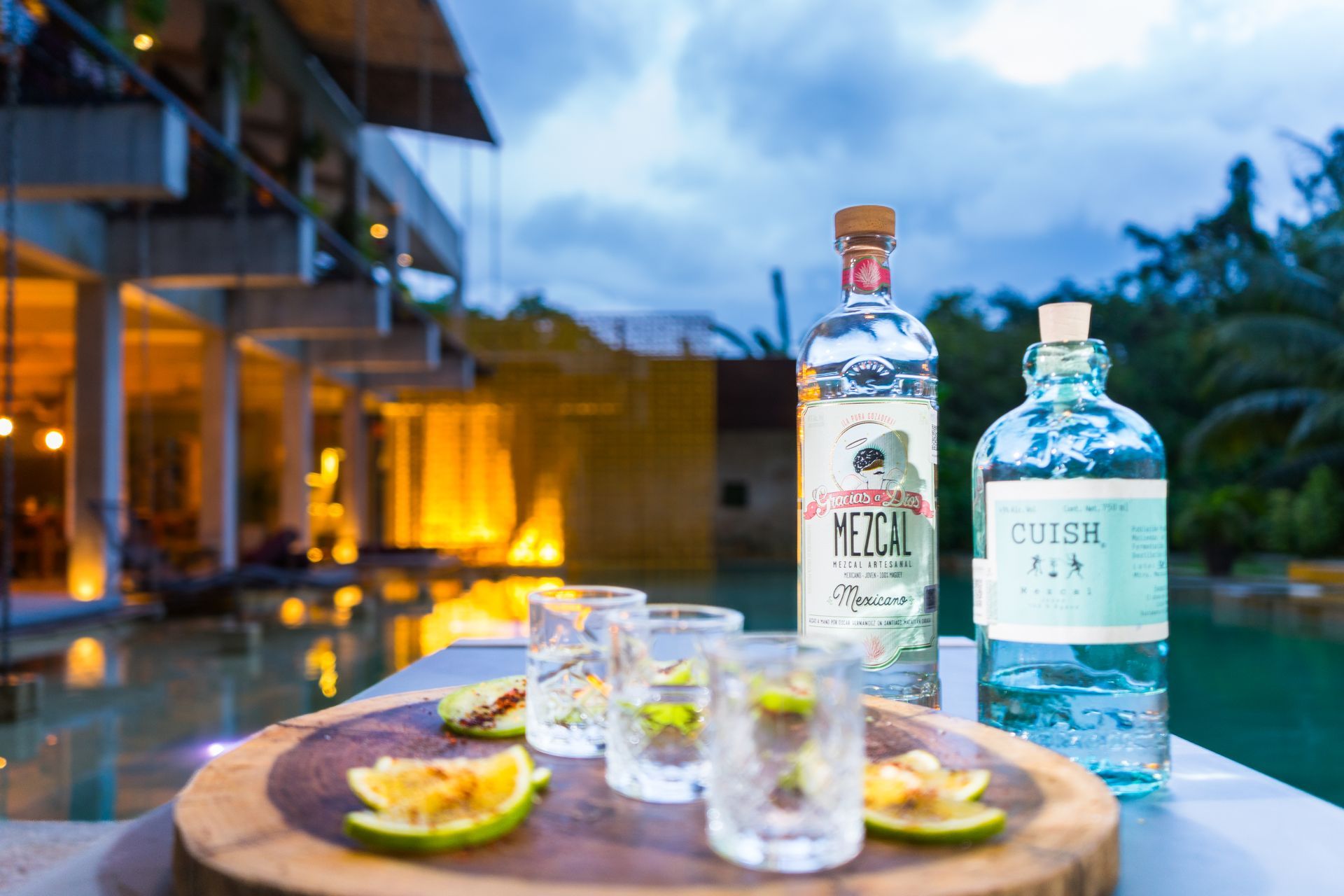 Botellas de tequila y ginebra con chupitos y rodajas de lima sobre una tabla de madera junto a una piscina. Fondo: edificio iluminado.
