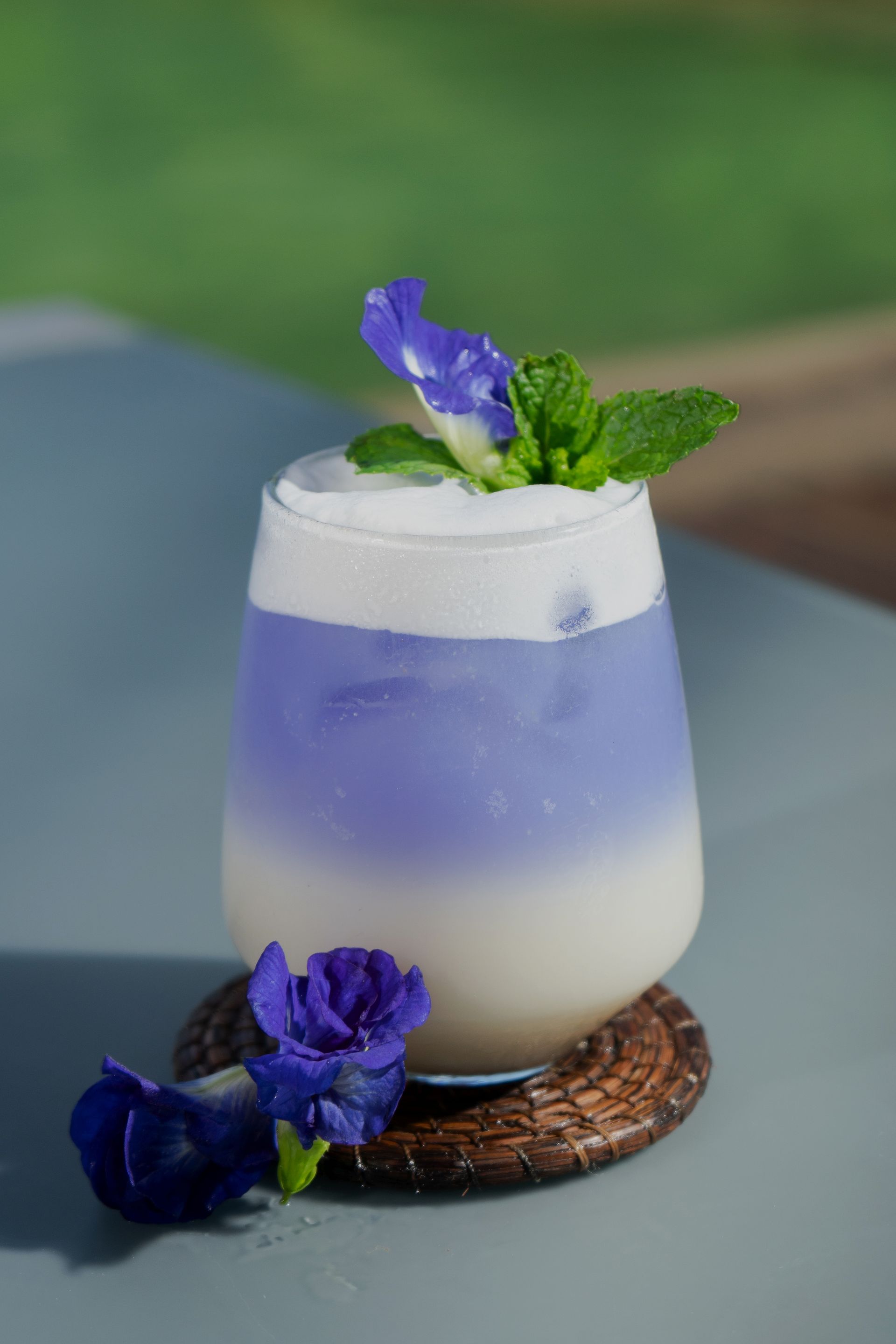 Bebida en capas de color morado y blanco adornada con flores y menta, colocada sobre un posavasos.