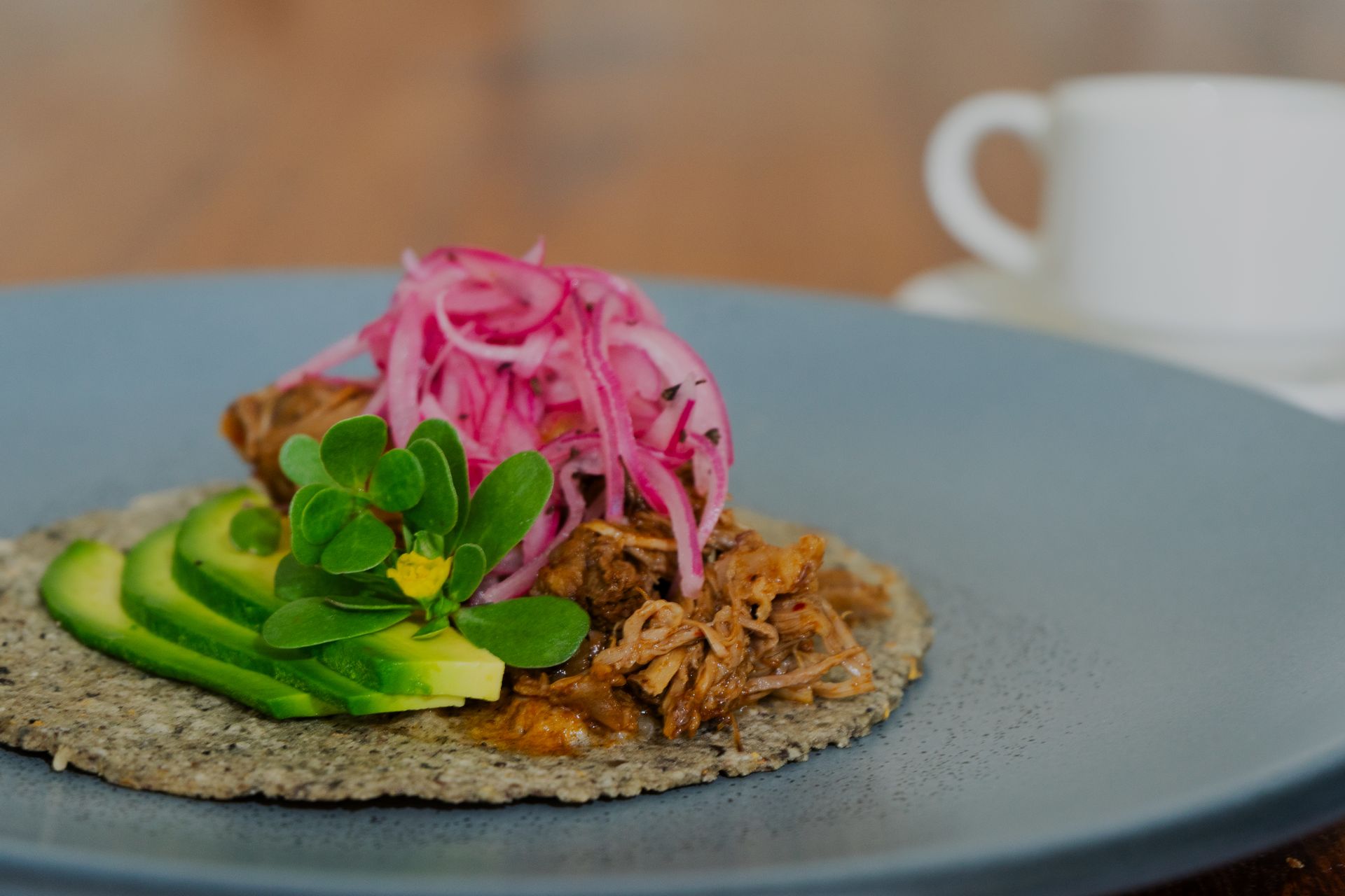 Taco con carne desmenuzada, cebollas encurtidas, rodajas de aguacate y una ramita de verduras en un plato azul; taza en el fondo.