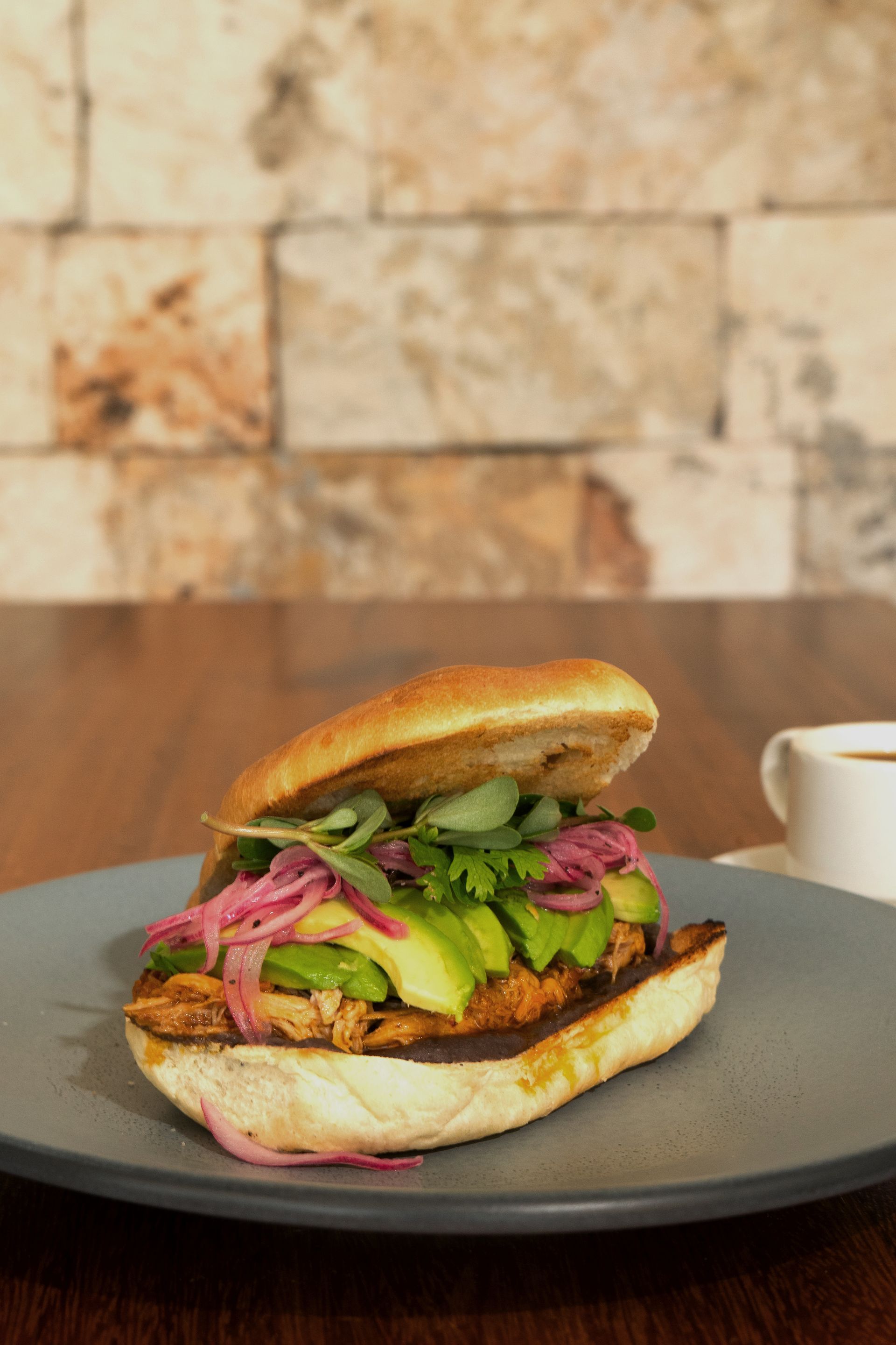 Sándwich con aguacate, cebolla encurtida y verduras en un plato gris; taza de café en el fondo.