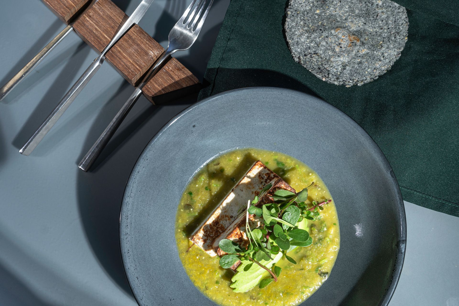 Sopa verde con guarnición en plato gris; cubiertos y decoración cerca.