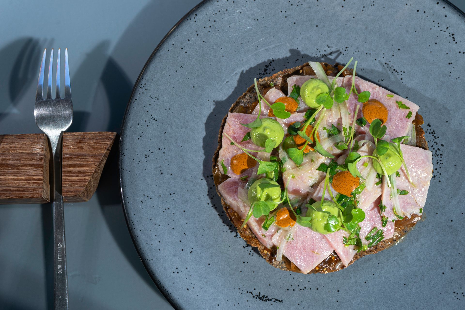 Tartar de atún sobre base de pan negro, cubierto con aguacate y salsa en un plato azul, con tenedor.