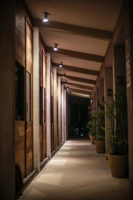 Largo pasillo exterior con puertas de madera y plantas en macetas, iluminado por focos.