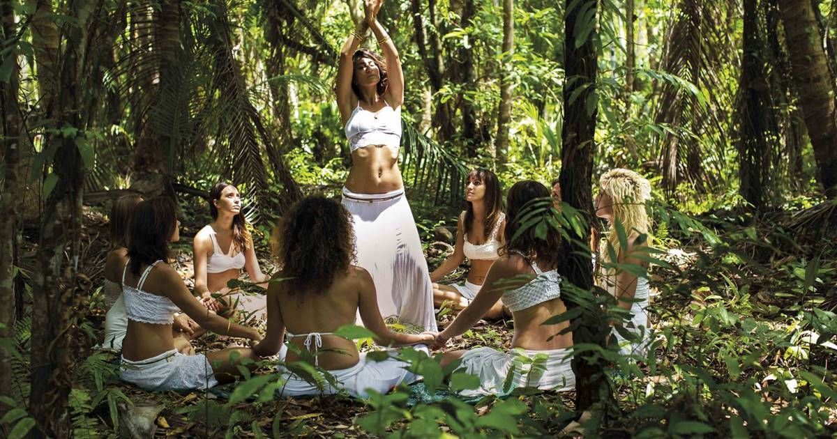 Grupo de mujeres vestidas de blanco forman un círculo, una de ellas con los brazos levantados, en un exuberante bosque verde.