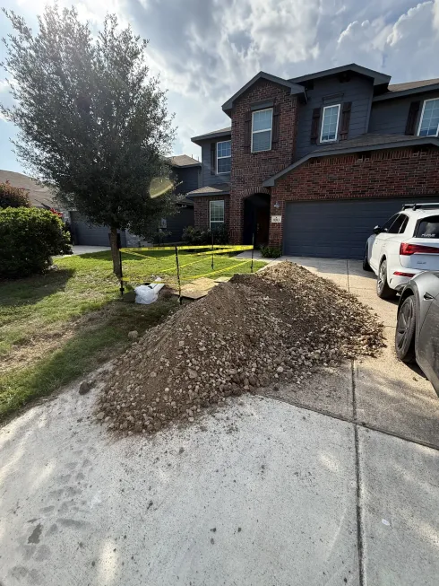 Pile of dirt on driveway beside a brick house and parked SUV under a cloudy sky