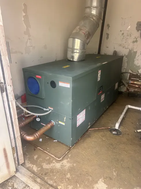 Green industrial furnace in a utility room with metal ductwork and exposed pipes.