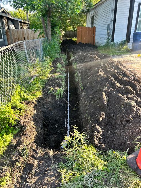 A water meter and pipe exposed in a dirt hole beside grass, with a hand nearby.