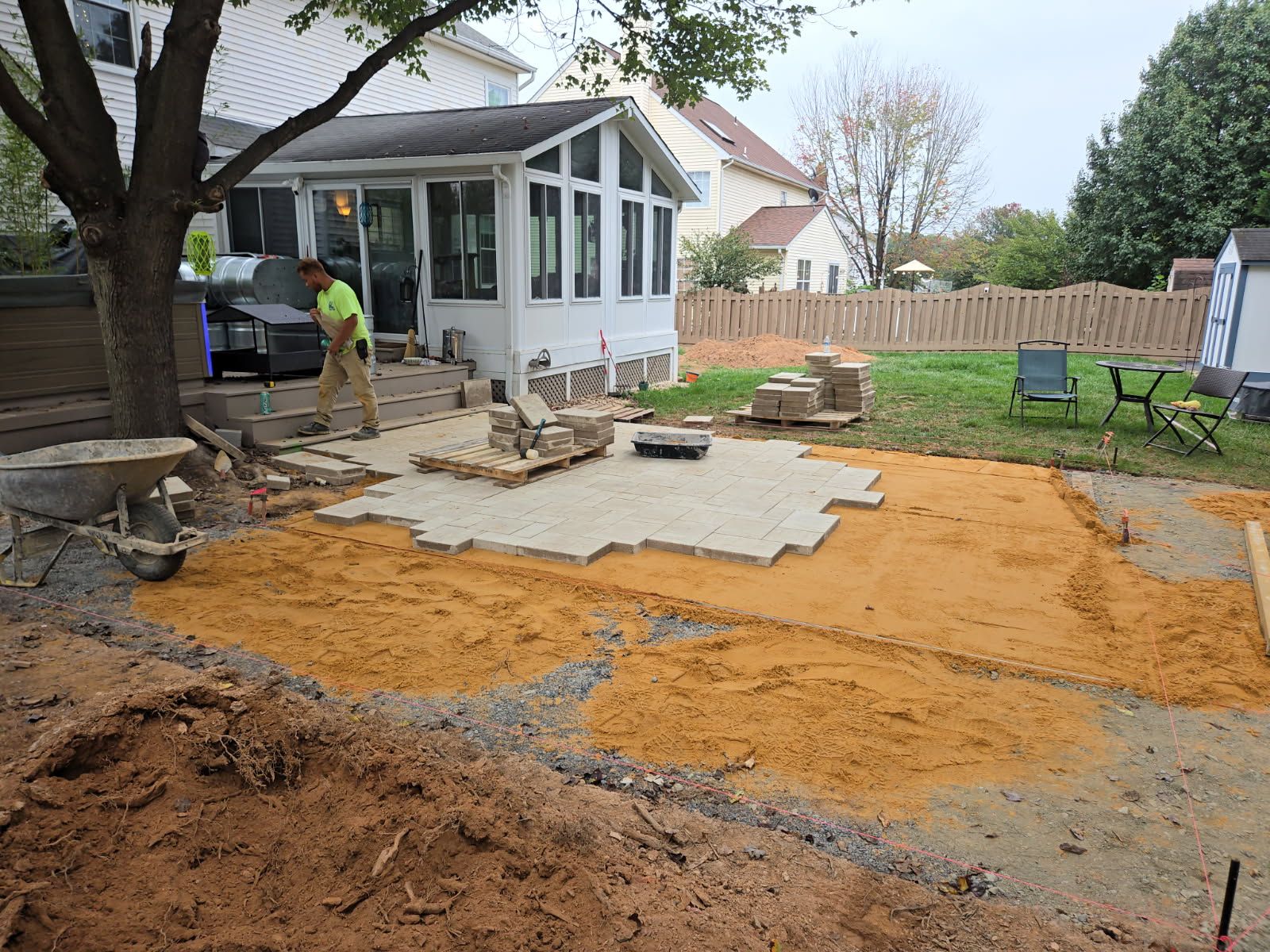 Backyard patio construction with pavers, sand, and a worker near a sunroom