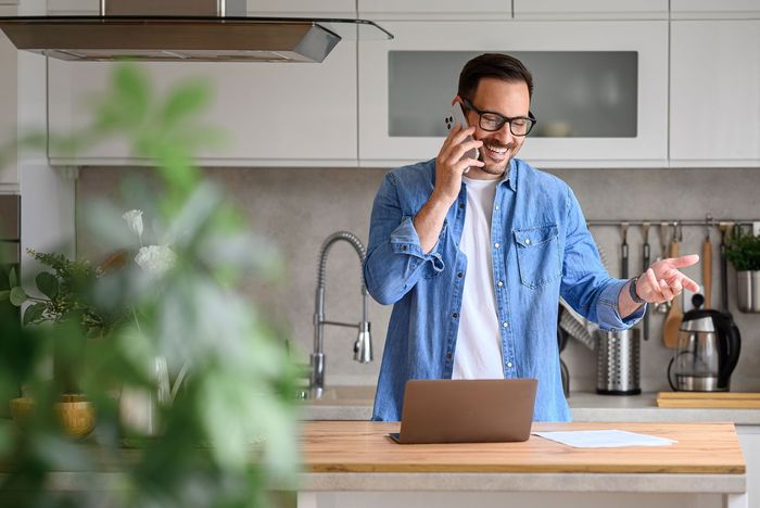 Person in a blue shirt on a phone call in a kitchen, standing by a laptop and coffee maker.