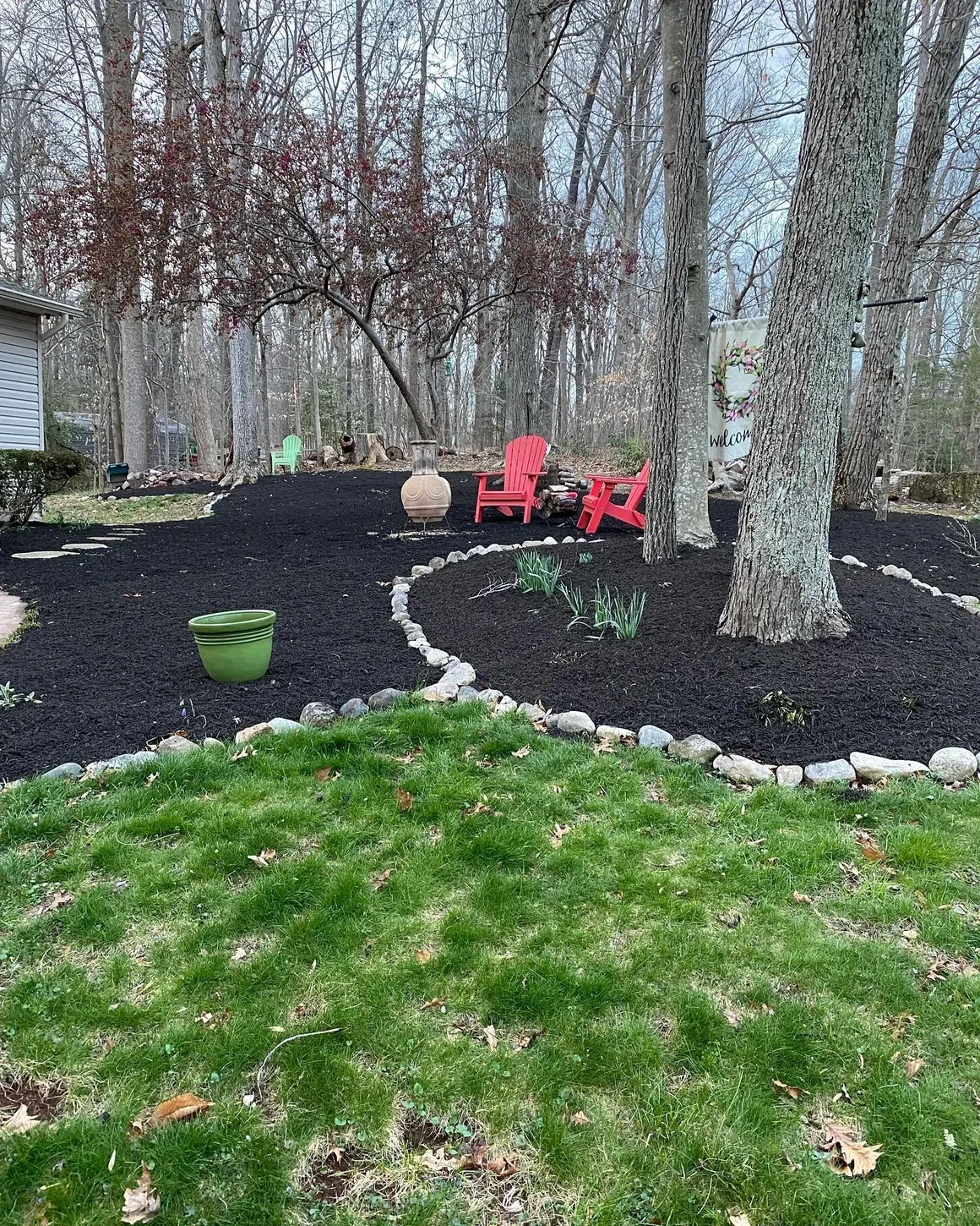 A backyard features a freshly mulched dark garden bed with stone borders, red Adirondack chairs, and a green planter.
