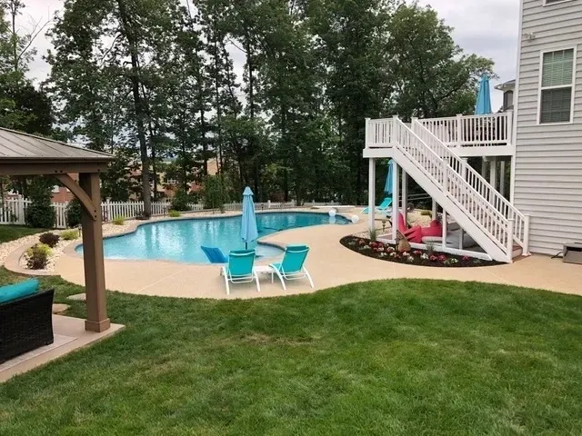 A backyard swimming pool with lounge chairs, a blue umbrella, a gazebo, and a raised wooden deck with white stairs.