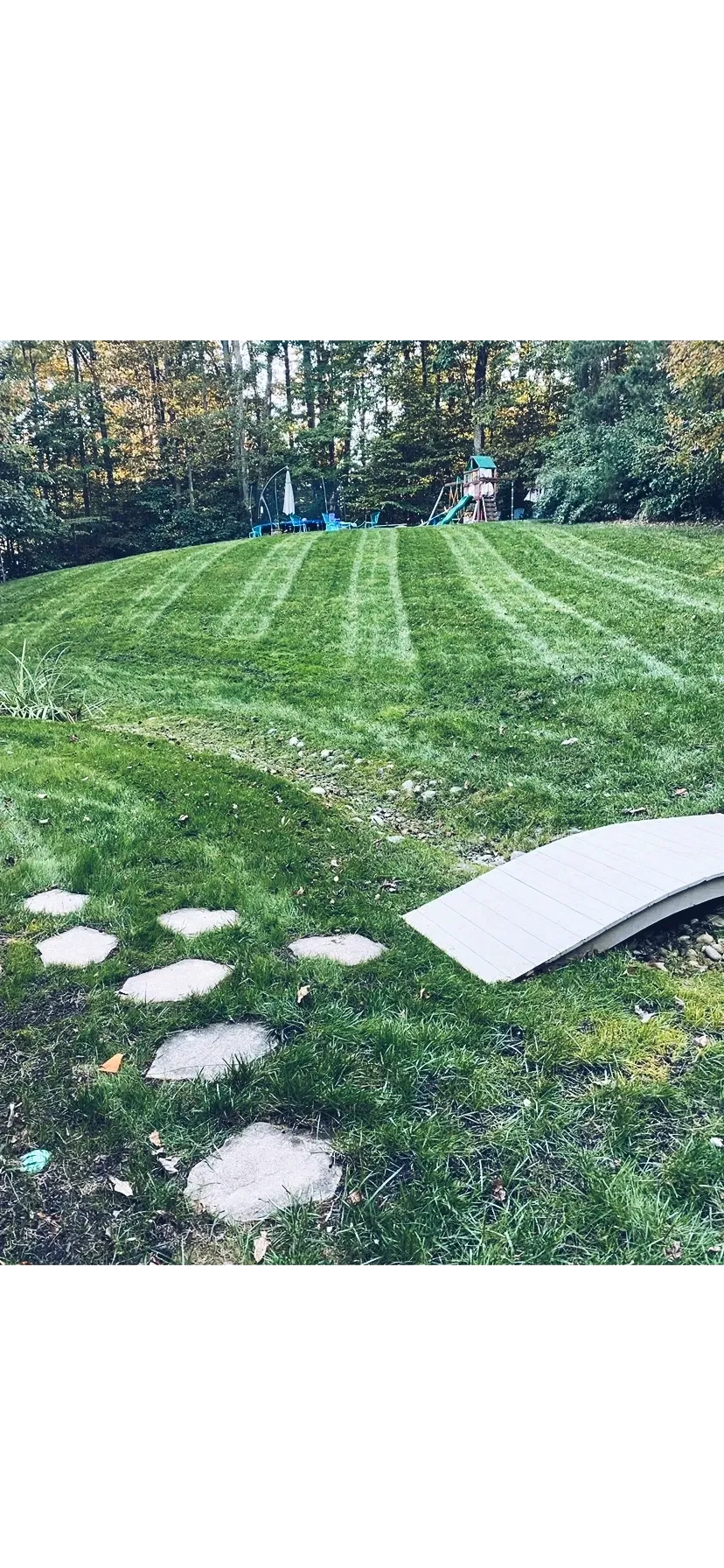 A lush green lawn with striped mowing patterns, leading to a small, arched wooden footbridge and stone stepping stones.