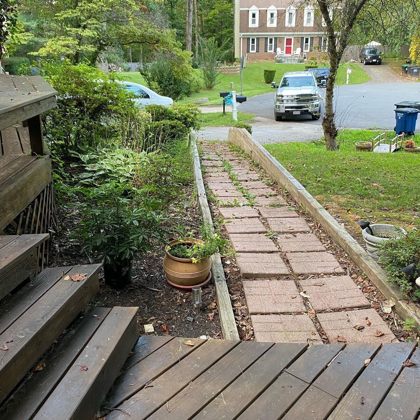 A view from a wooden deck looking out toward a brick pathway leading through a residential yard with a house in the back.
