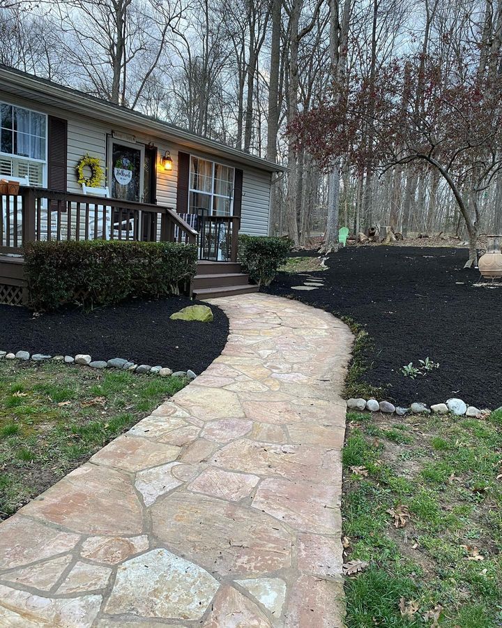 A stone walkway leads to the front porch of a light-colored house with dark shutters, surrounded by black mulch and trees.