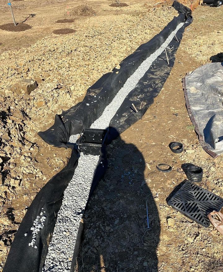 A French drain under construction with a gravel-filled trench, landscape fabric, and a catch basin in a dirt yard.