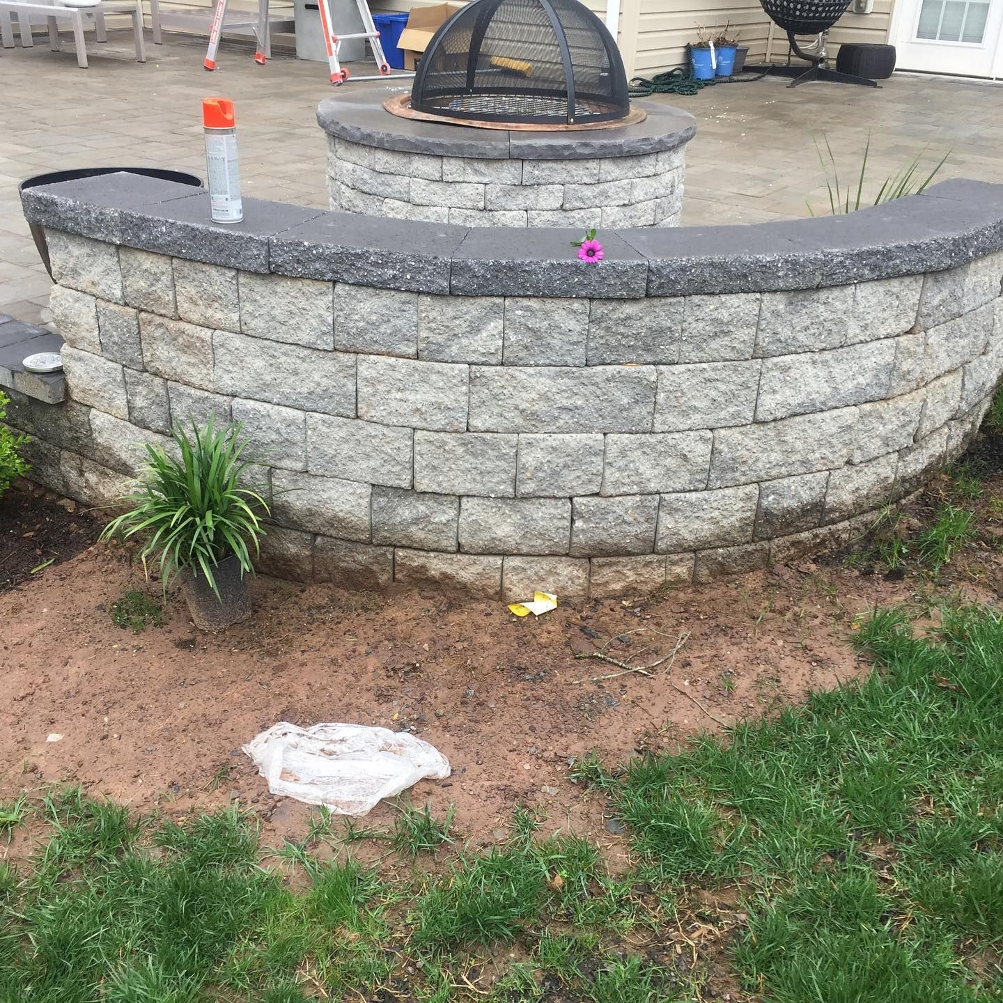 A curved, stone retaining wall with a fire pit behind it on a patio, with a small potted plant and debris on the ground.
