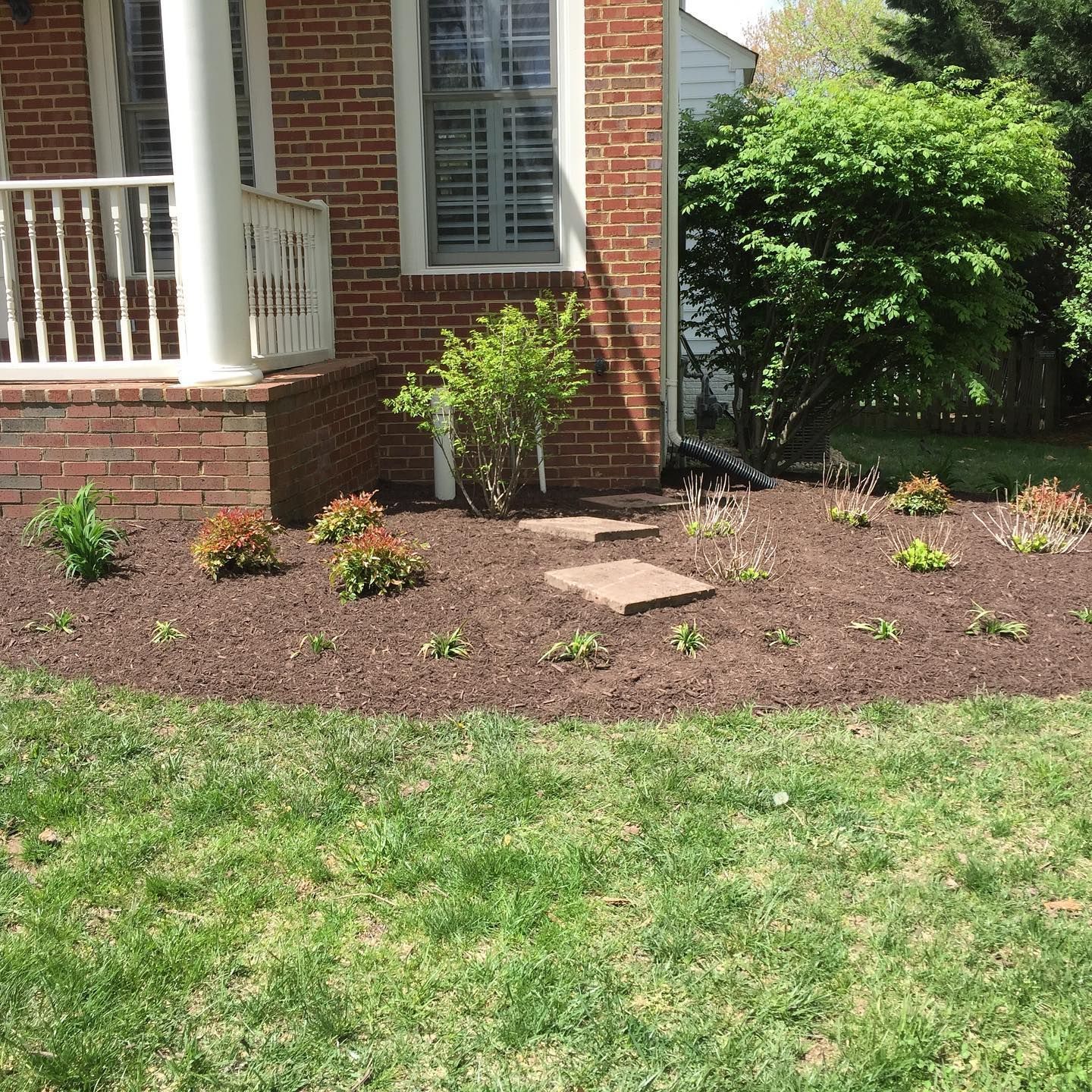 A brick house foundation featuring a porch, a small shrub, and a garden bed covered in dark mulch with small plants.