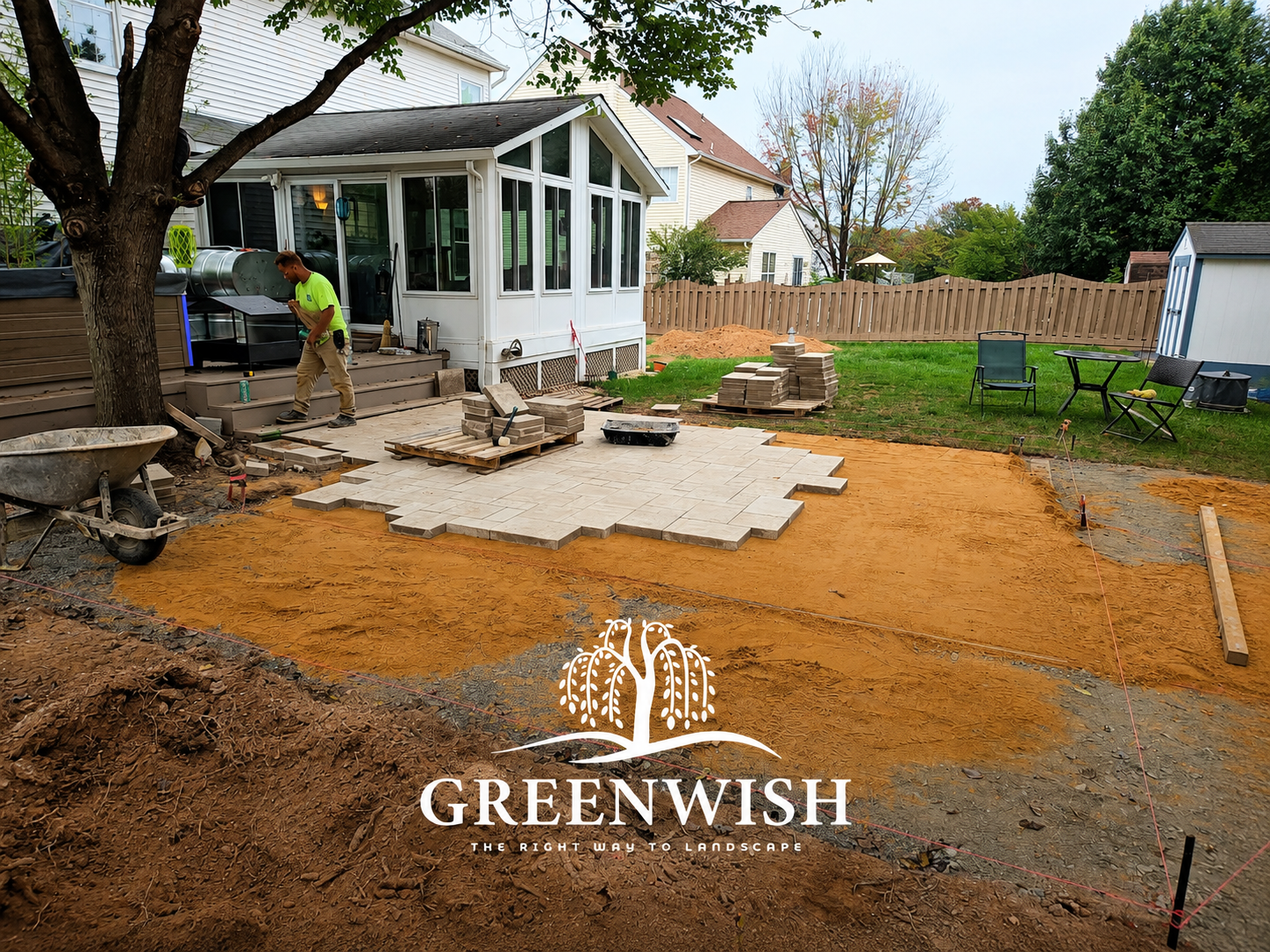 A worker wearing work clothes and knee pads installs rectangular gray stone pavers on a gravel bed.