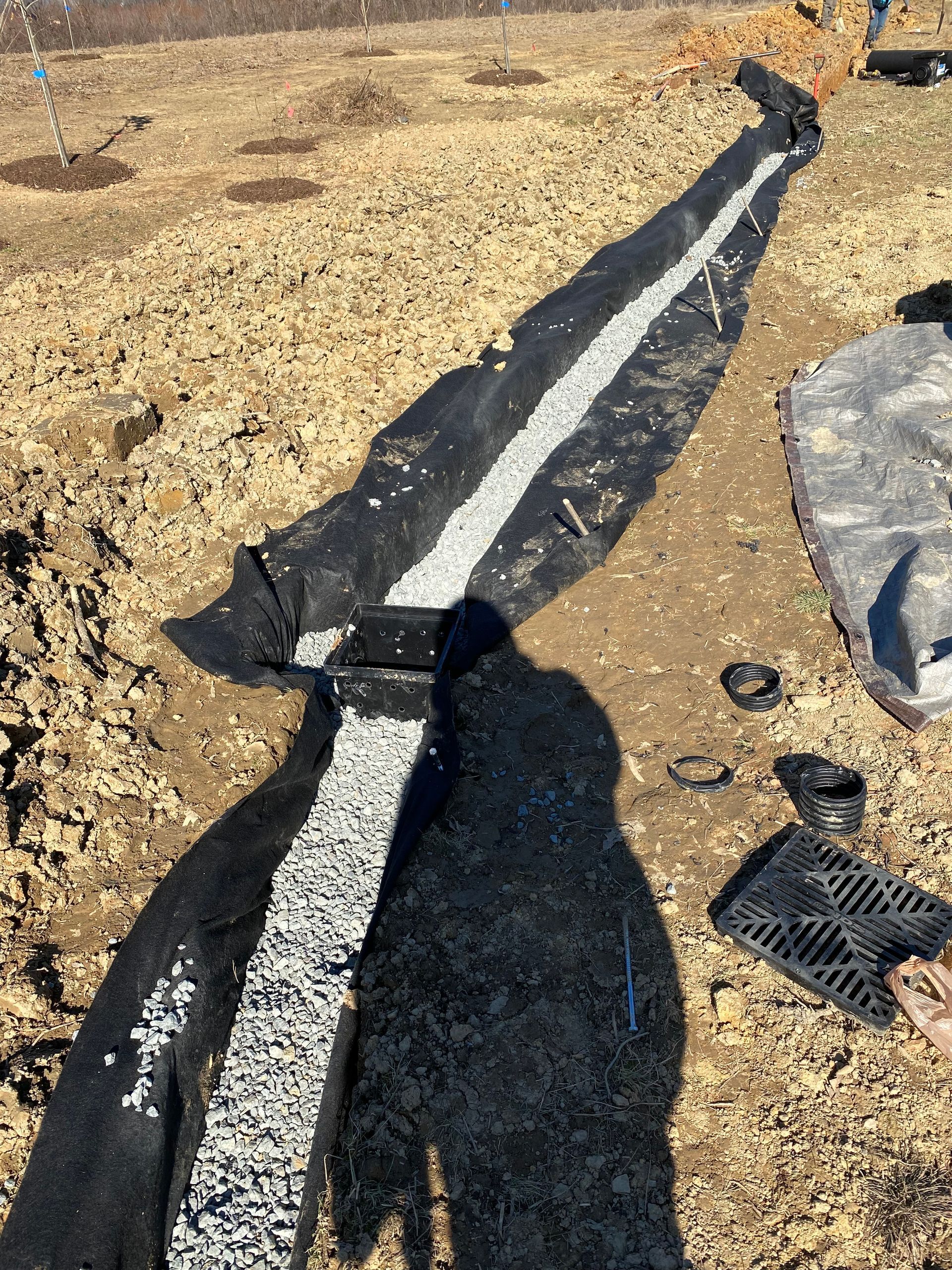 A drainage trench lined with black landscape fabric and filled with white gravel, with a catch basin grate on the side.
