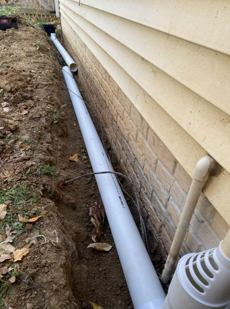 White drainage pipe runs along a house foundation in a narrow dirt trench.