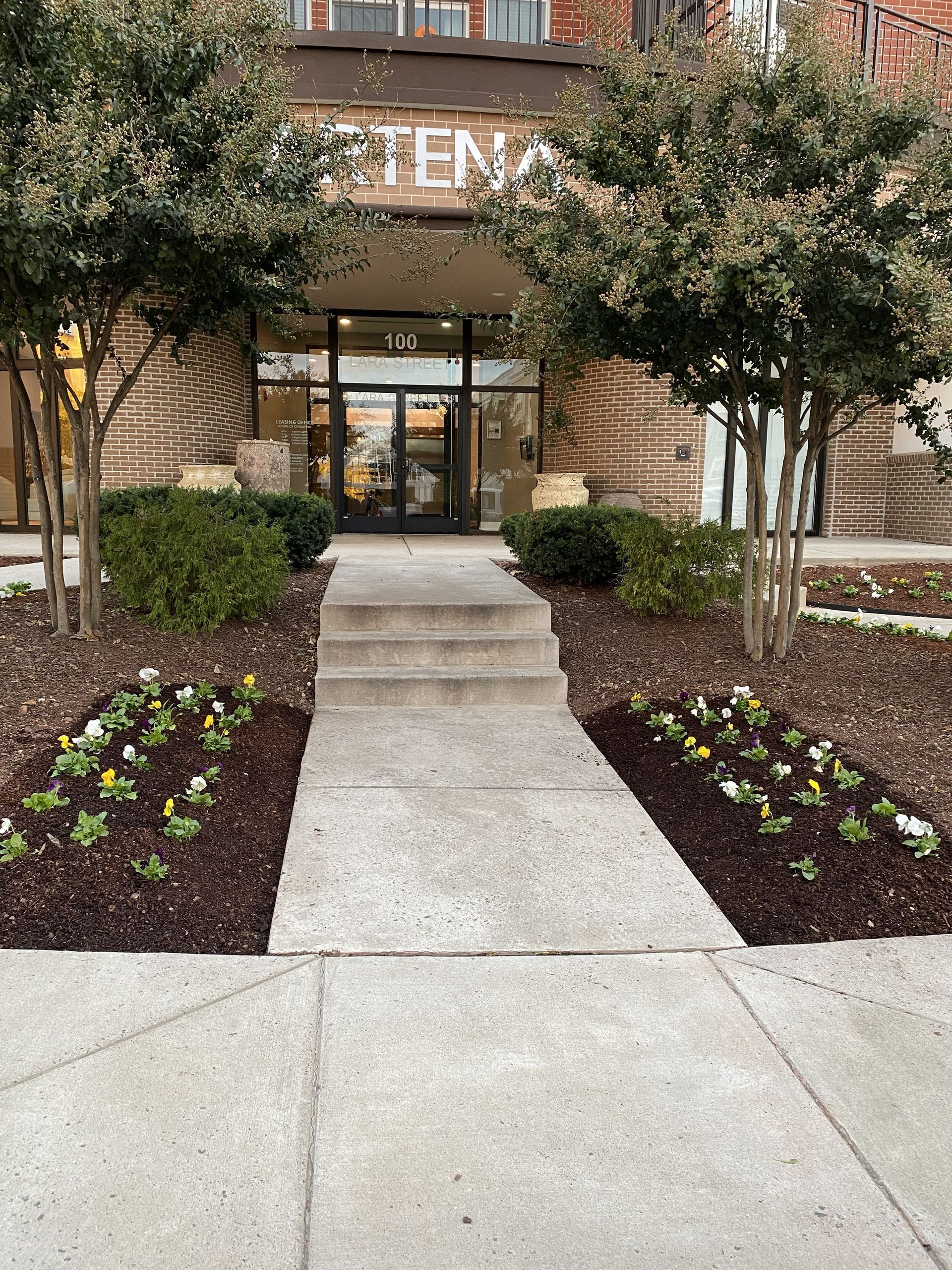 A brick building entrance at 133, featuring a concrete walkway flanked by mulched garden beds with small flowers and trees.