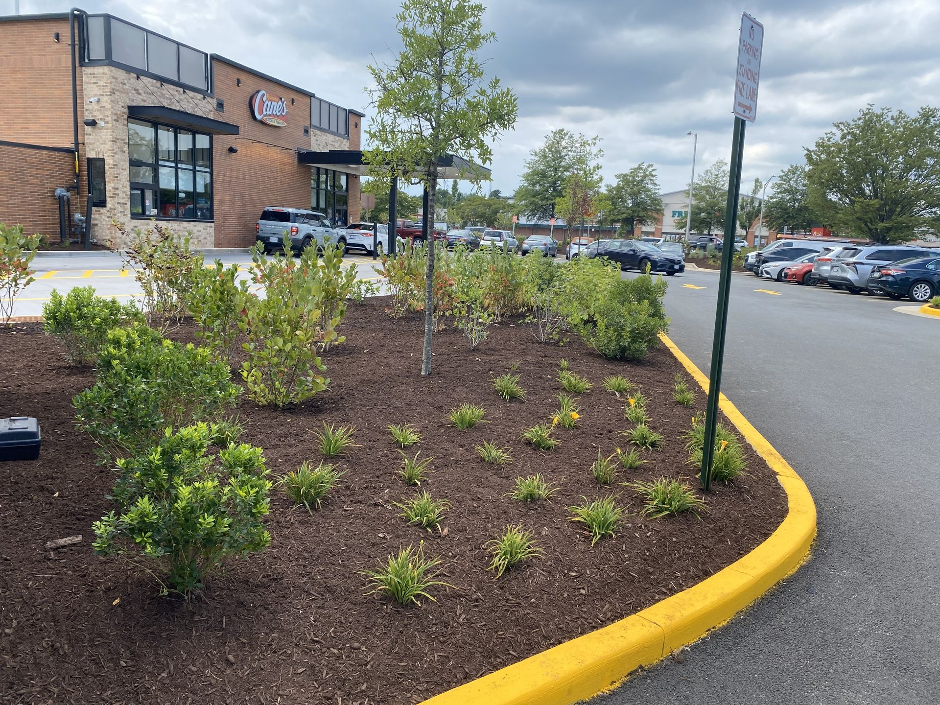 Landscaped parking lot with shrubs, yellow curb, and a brick building under a partly cloudy sky