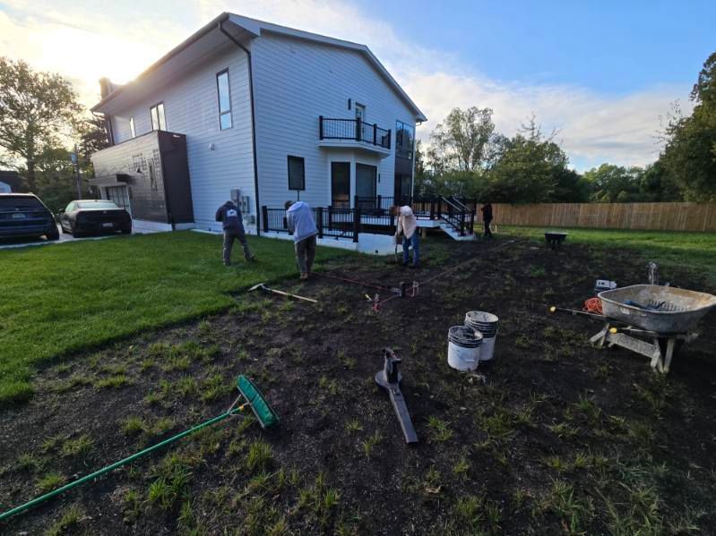 Three people work on landscaping a backyard, preparing the soil near a multi-story house and deck.