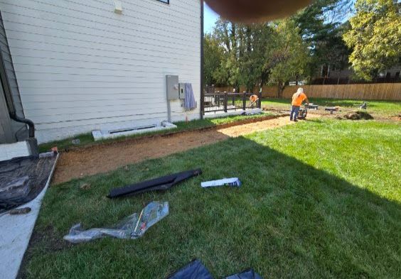 A worker operates machinery on a dirt path under construction next to a white house with a green lawn.