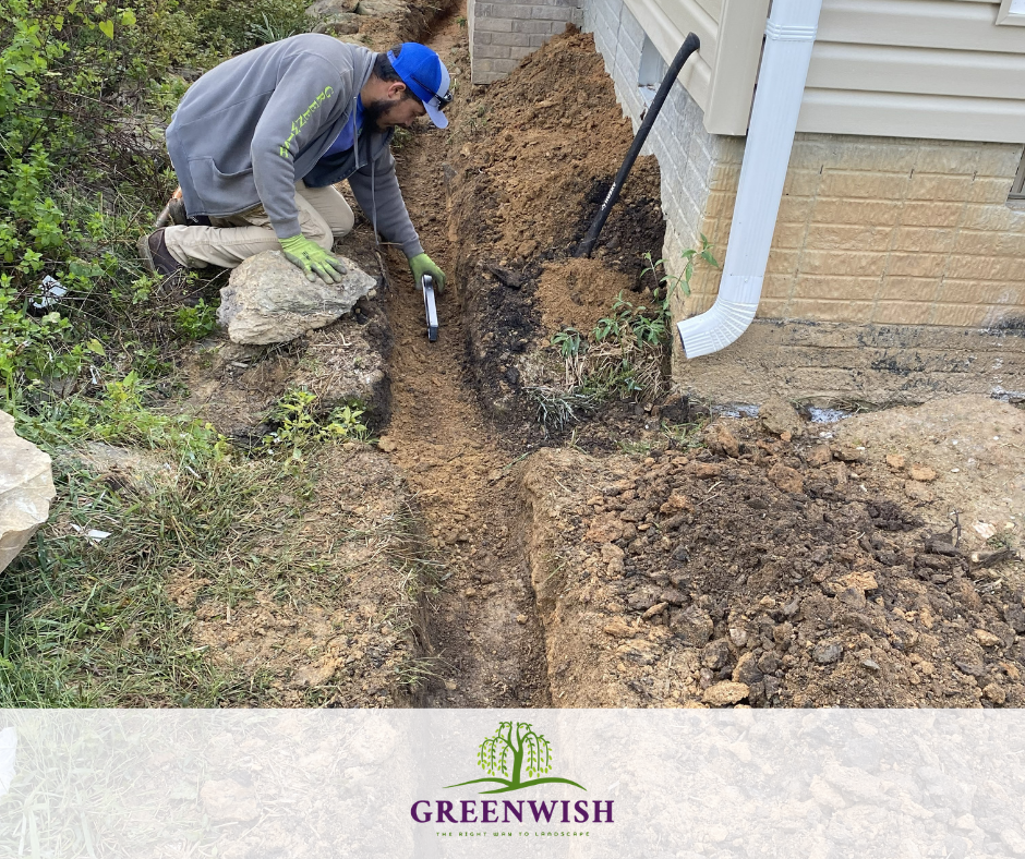 Worker digs a trench beside a house with a shovel and hose, clearing soil along the foundation.