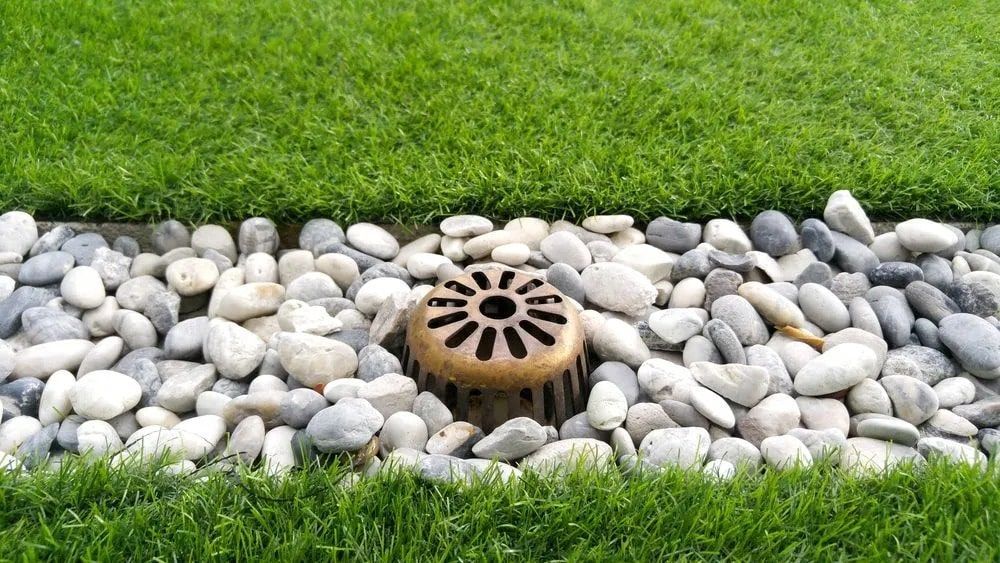 A brass-colored dome-shaped drain cover sits in a trench filled with smooth, multicolored river stones between lawn grass.