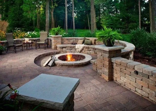 A stone patio featuring a sunken fire pit surrounded by a curved stone wall with seating, plants, and chairs.