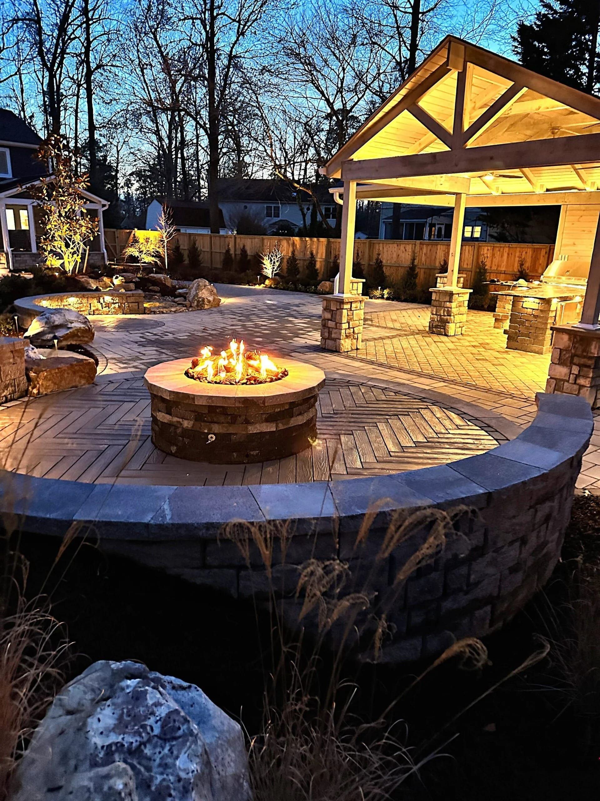 A cozy backyard patio at twilight with a glowing fire pit, stone seating, and an illuminated timber-framed pavilion.