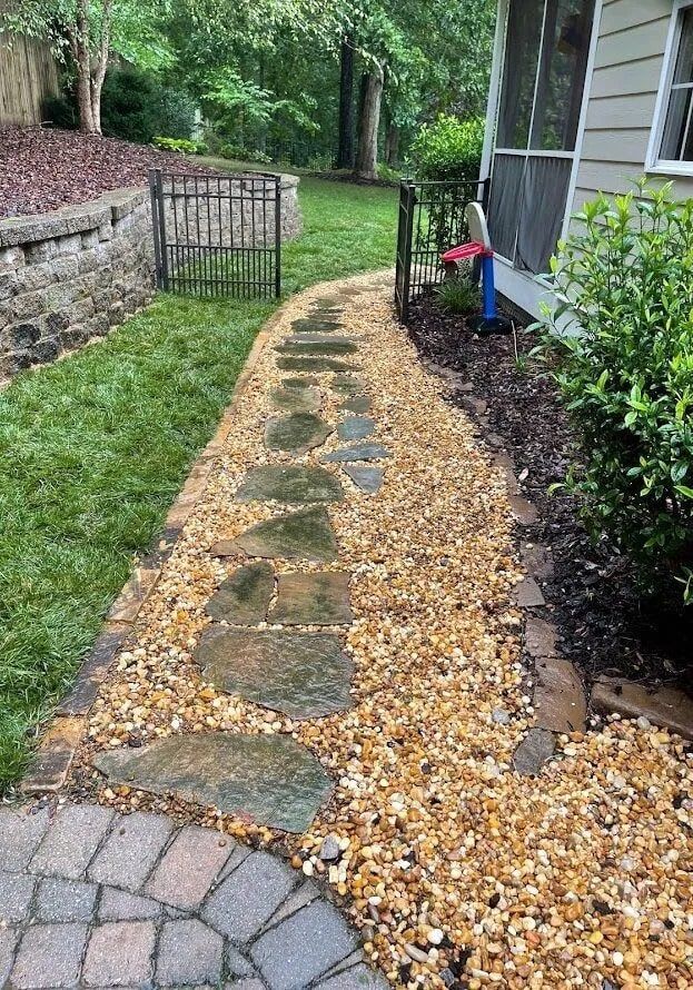 A stone pathway lined with golden gravel leading along the side of a house toward a small metal gate in a grassy yard.