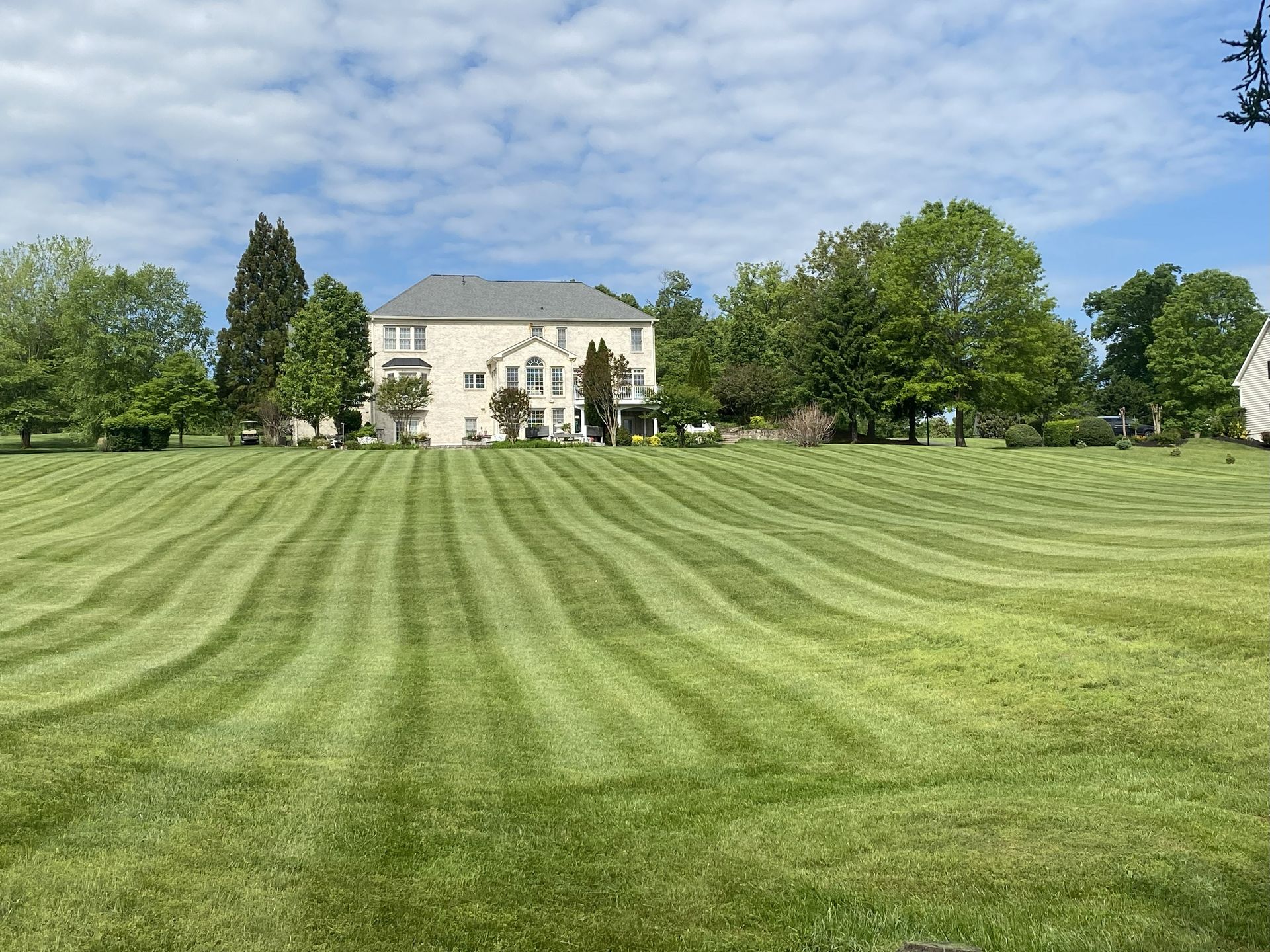 A large beige house sits in the background of a vast, sunny lawn featuring meticulously mowed, wavy grass stripes.