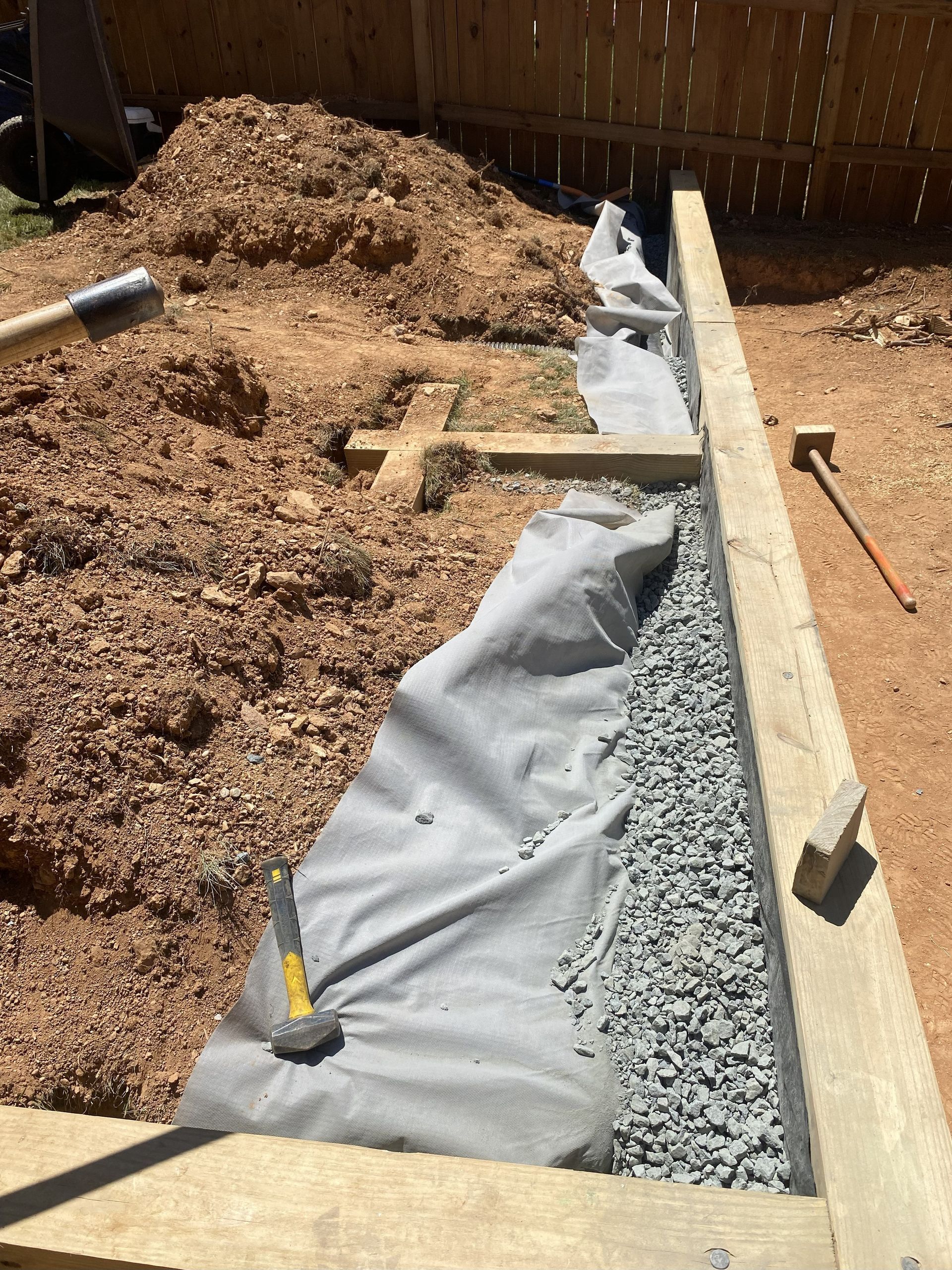 An outdoor project site featuring a wooden retaining wall frame, gravel drainage trench, and landscaping fabric.