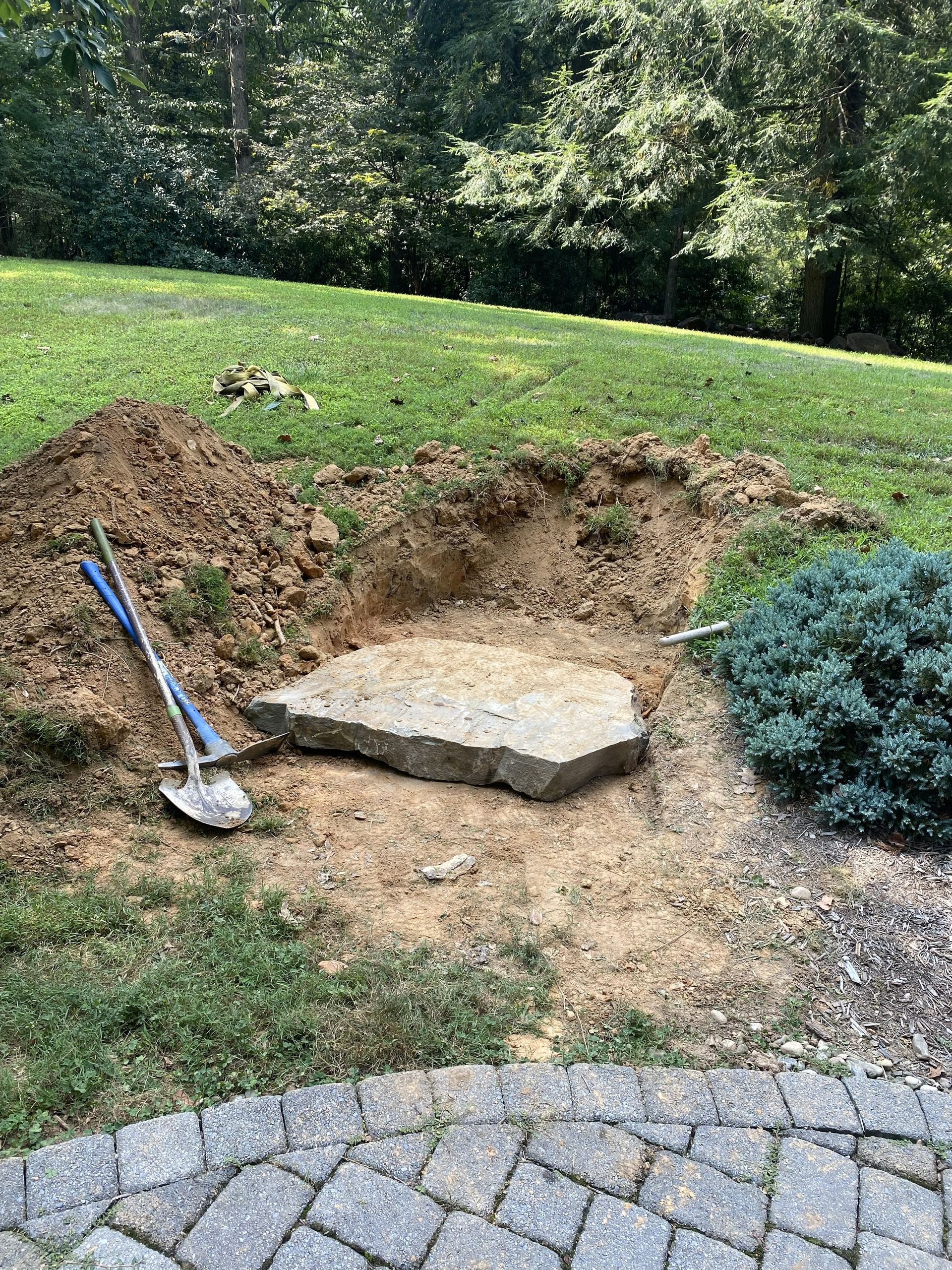 A large, flat stone sits in a freshly dug hole in a lawn, next to a shovel and a pile of dirt near a stone-paved patio.